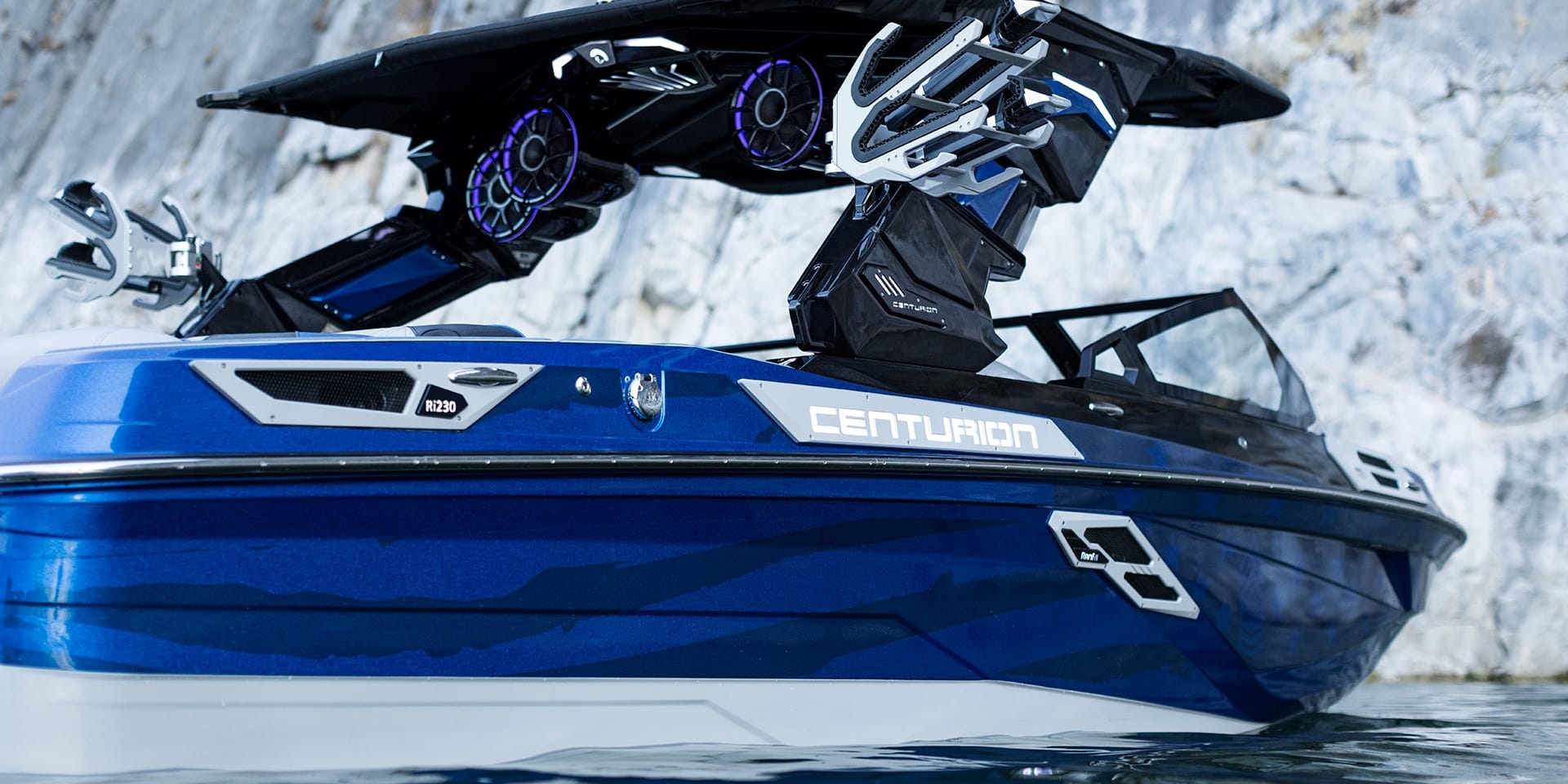 A blue Centurion Ri230 with large speakers is docked near a rocky shore.