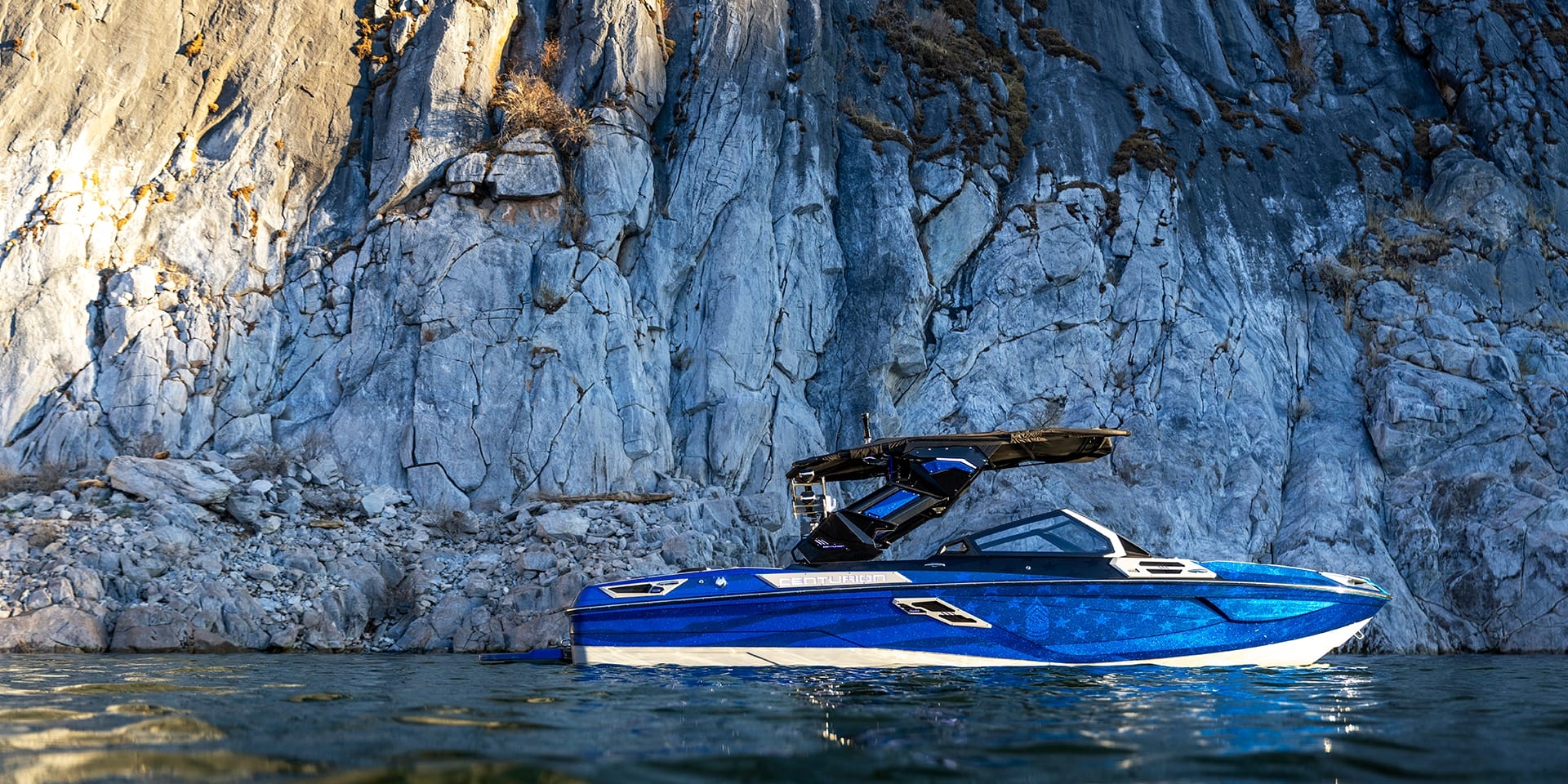 A blue Centurion Ri230 boat is gracefully floating on the water in front of a large rocky cliff.