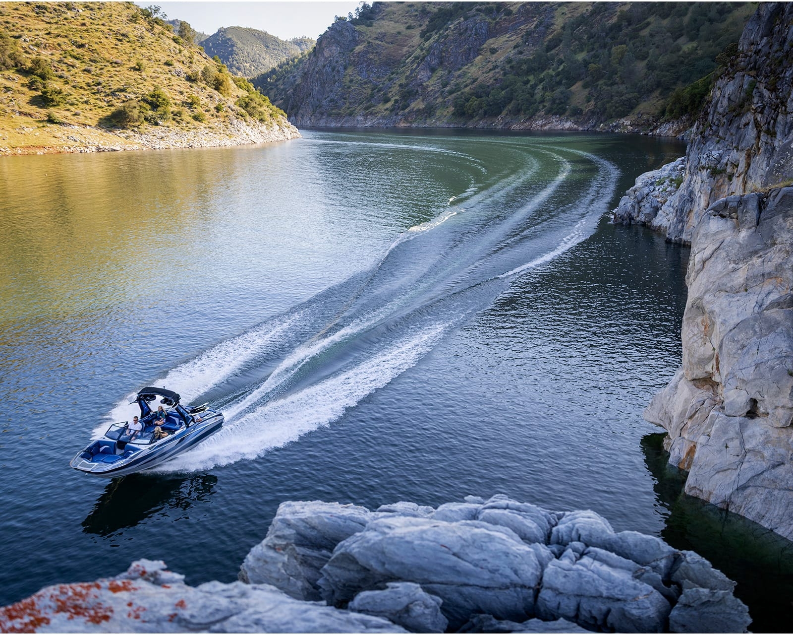 A Centurion Fi23 surfs along a river, leaving a foamy wake behind as it passes near rocky cliffs and green hills under a clear sky.
