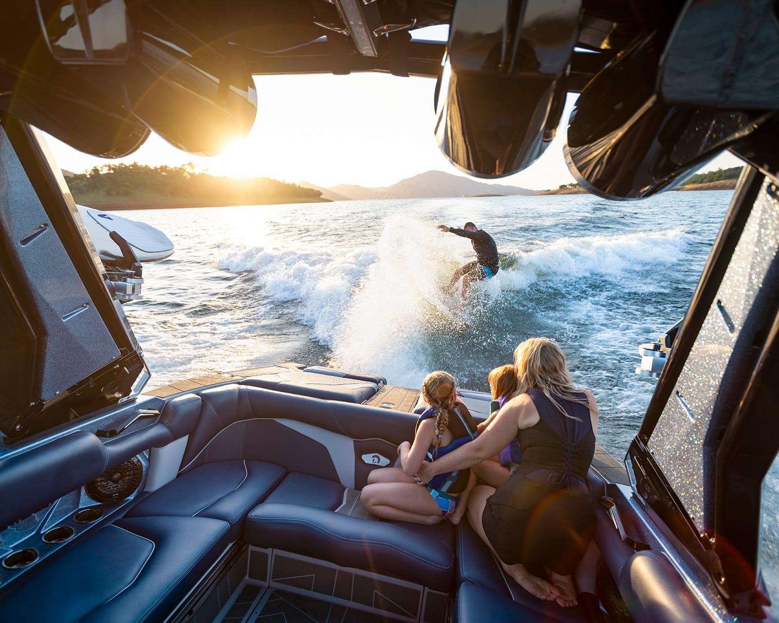 A person wake surfs behind a Centurion Fi23 near a lake shore at sunset, while three people watch from the boat's open seating area.