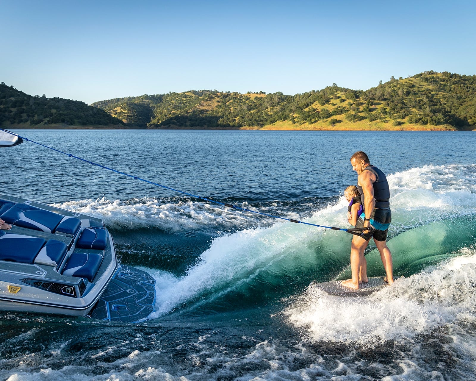 A man and a young girl wakeboard together behind a Centurion Fi23 on a lake, while a woman on the boat watches and takes photos; hills and trees are visible in the background.