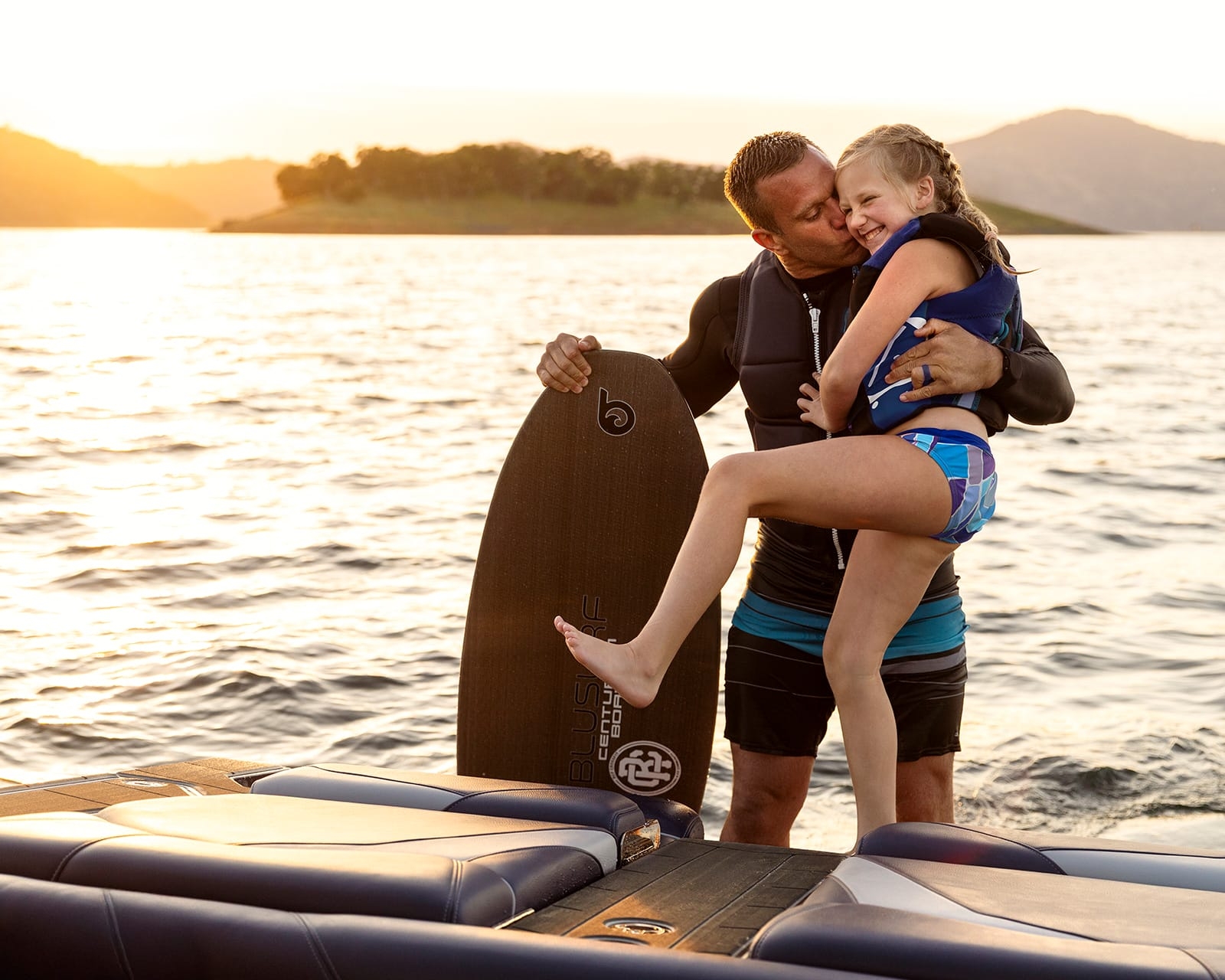 A man in a wetsuit holding a wakeboard hugs and lifts a smiling girl in a swimsuit by a lake at sunset, with their Centurion Fi23 surf boat gleaming on the water behind them.