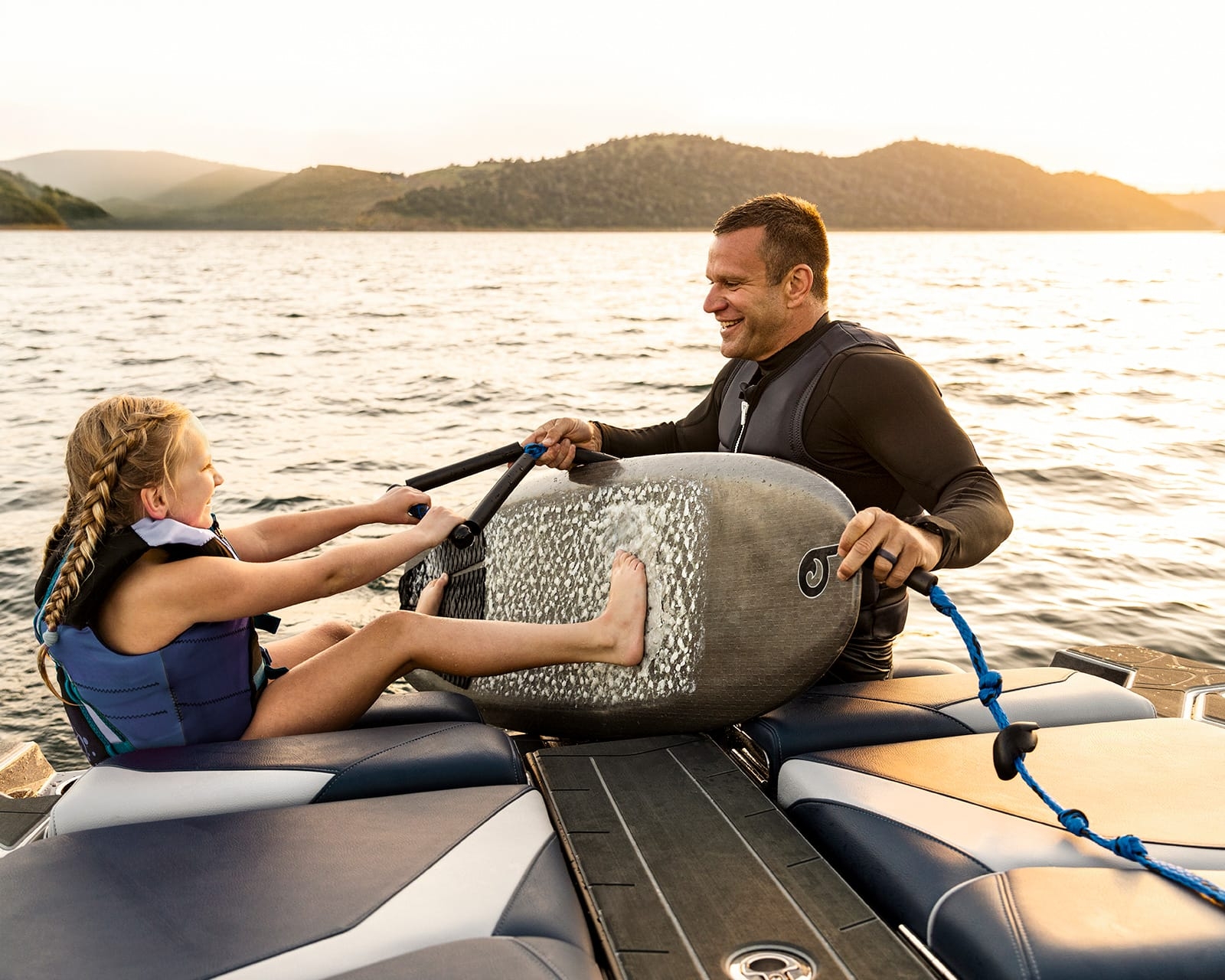 A man and a young girl wearing life jackets sit on a docked Centurion Fi23 at sunset, holding onto a wakesurf board together and smiling.