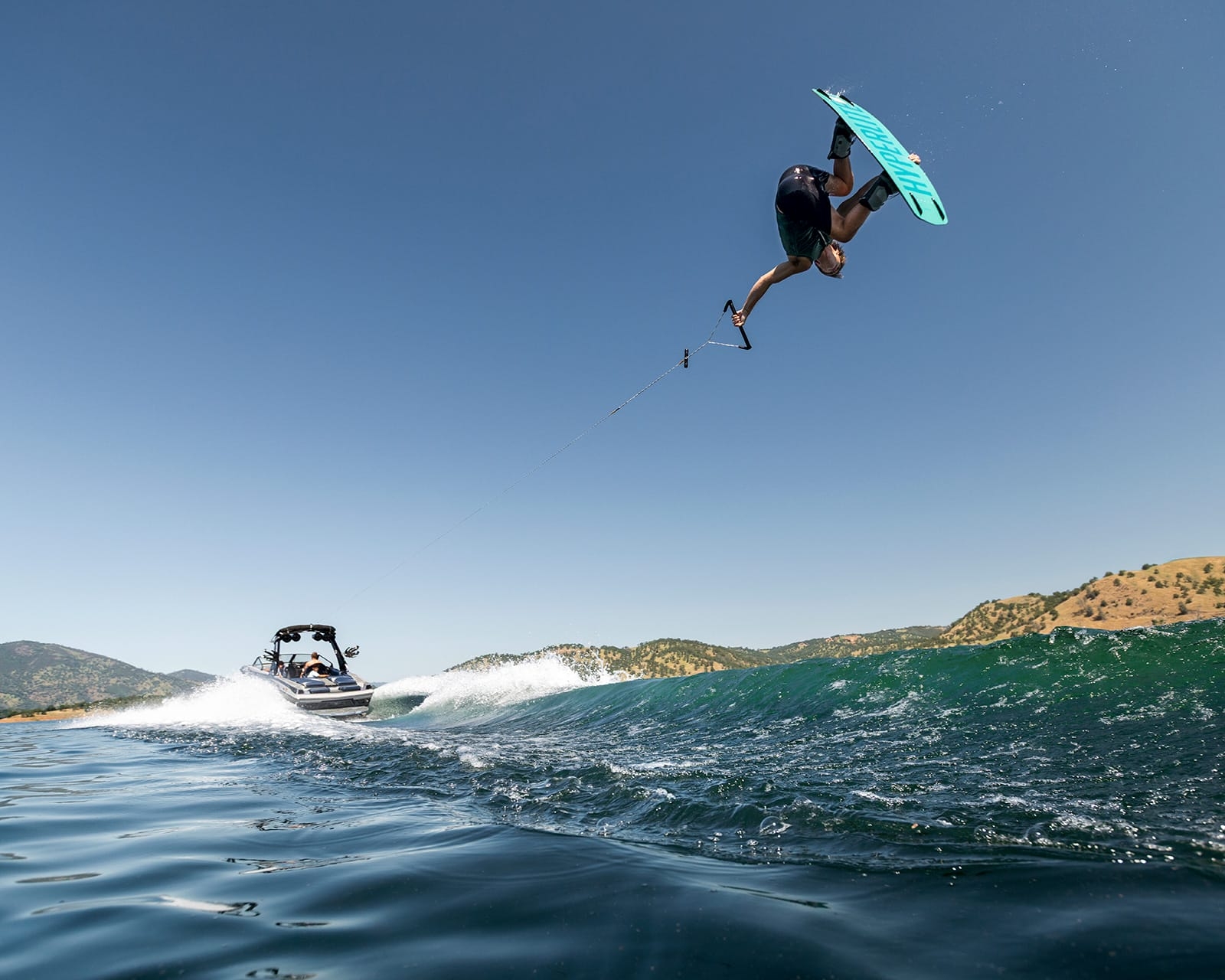 A person wakeboarding performs a high jump trick over the water, holding a rope attached to a Centurion Fi23, with hills and a clear sky in the background.