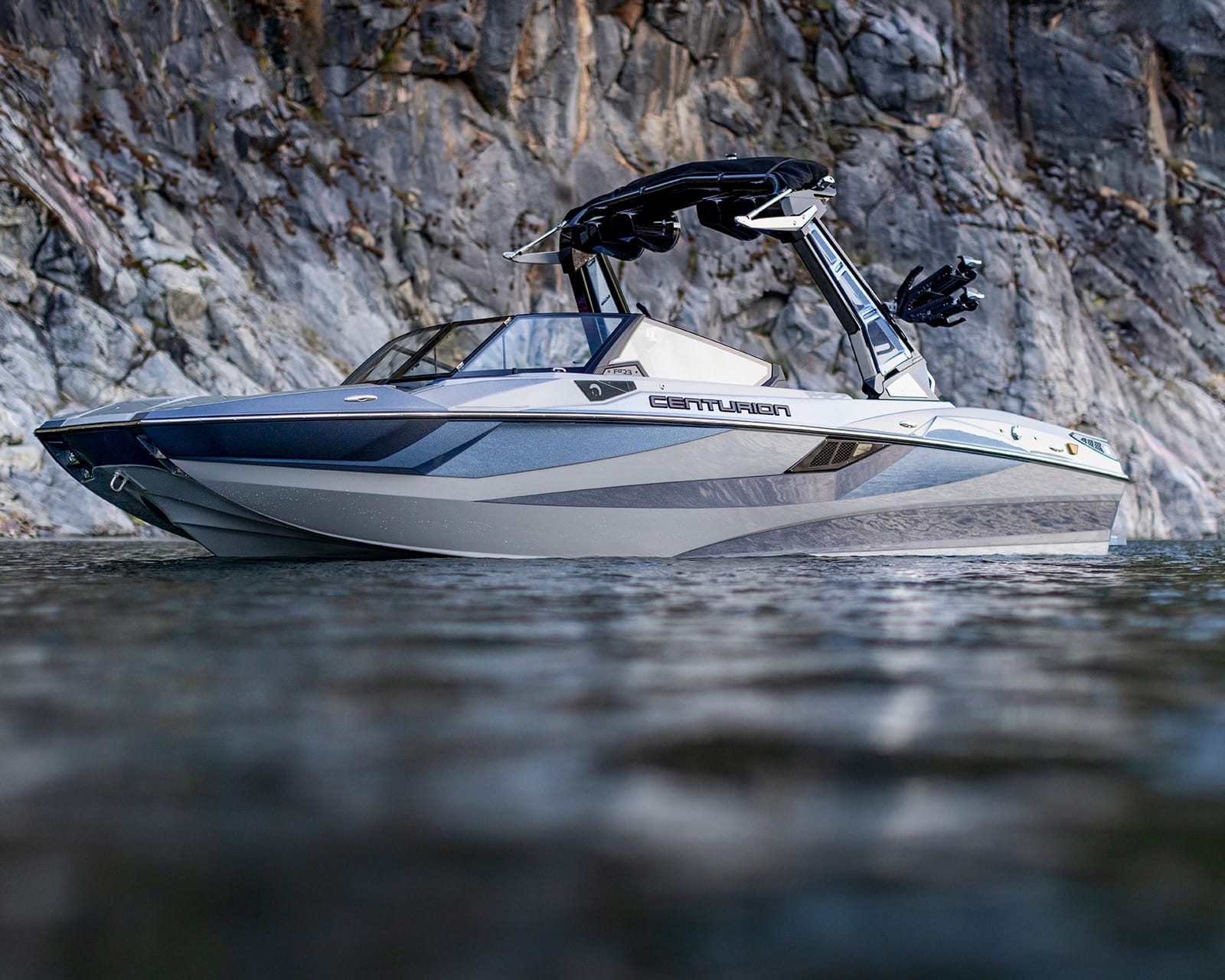 A modern Centurion Fi23 with a wakeboard tower is floating on calm water near a rocky shoreline.