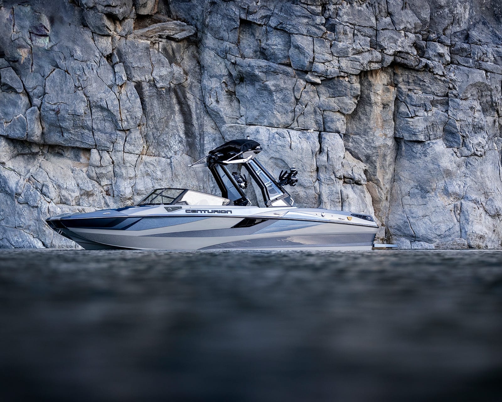 A white and gray Centurion Fi23 speedboat floats on calm water near a large rocky cliff.