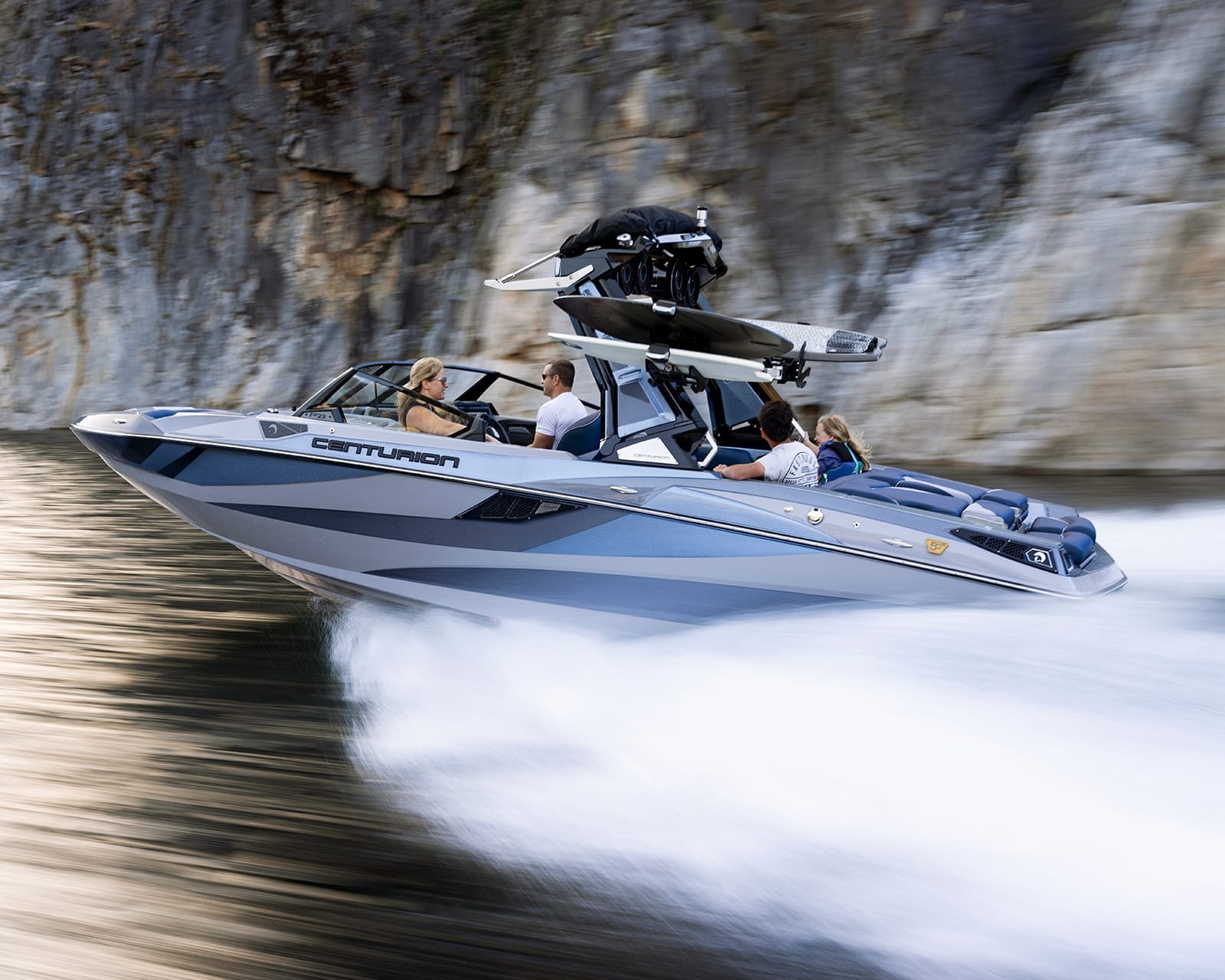 A Centurion Fi23 speedboat with four people moves quickly across the water near a rocky shoreline, leaving a trail of white spray behind.
