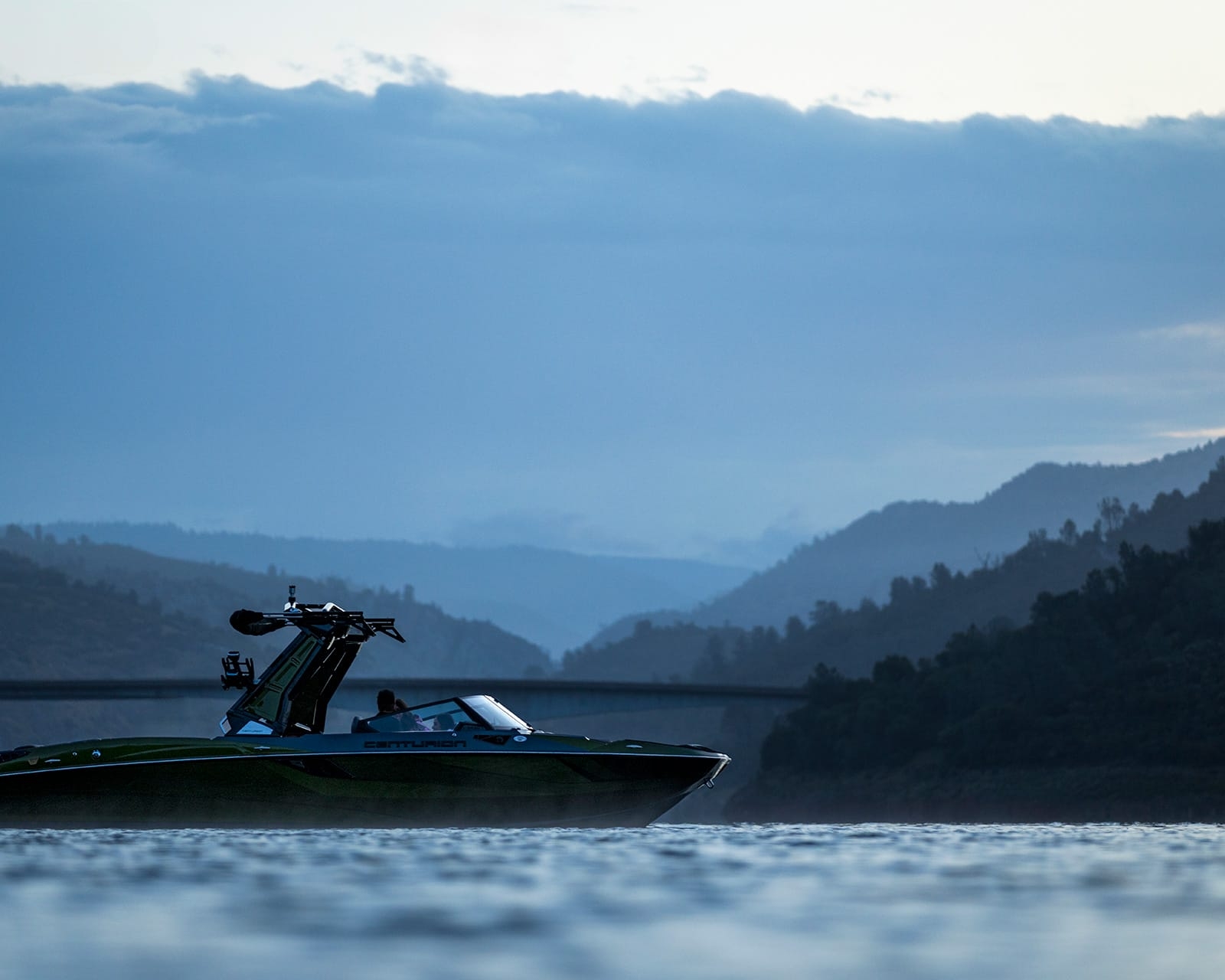 A Centurion Fi25 glides on the water with layered hills and a bridge in the background, all under a cloudy sky at dusk.