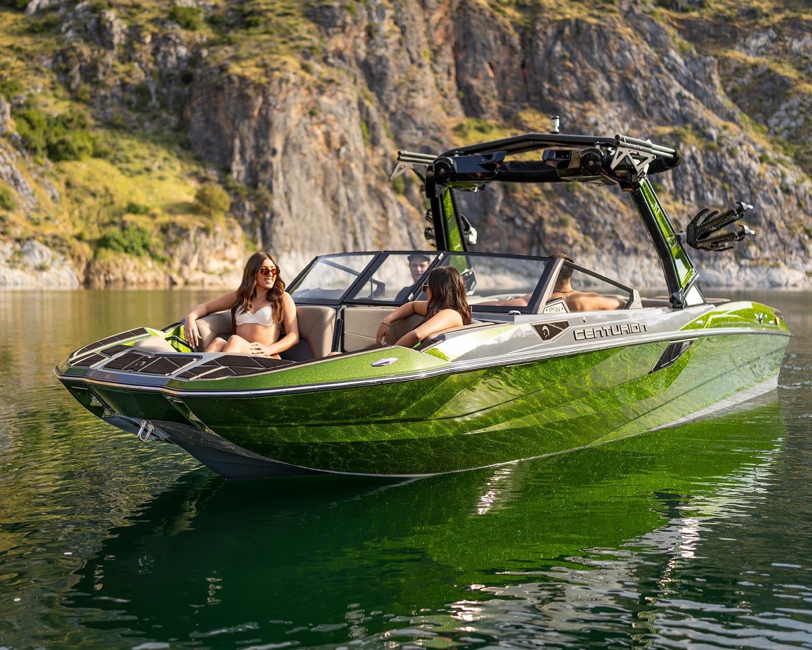Two women sit on a green Centurion Fi25 surf boat floating on calm water near rocky cliffs and lush greenery.