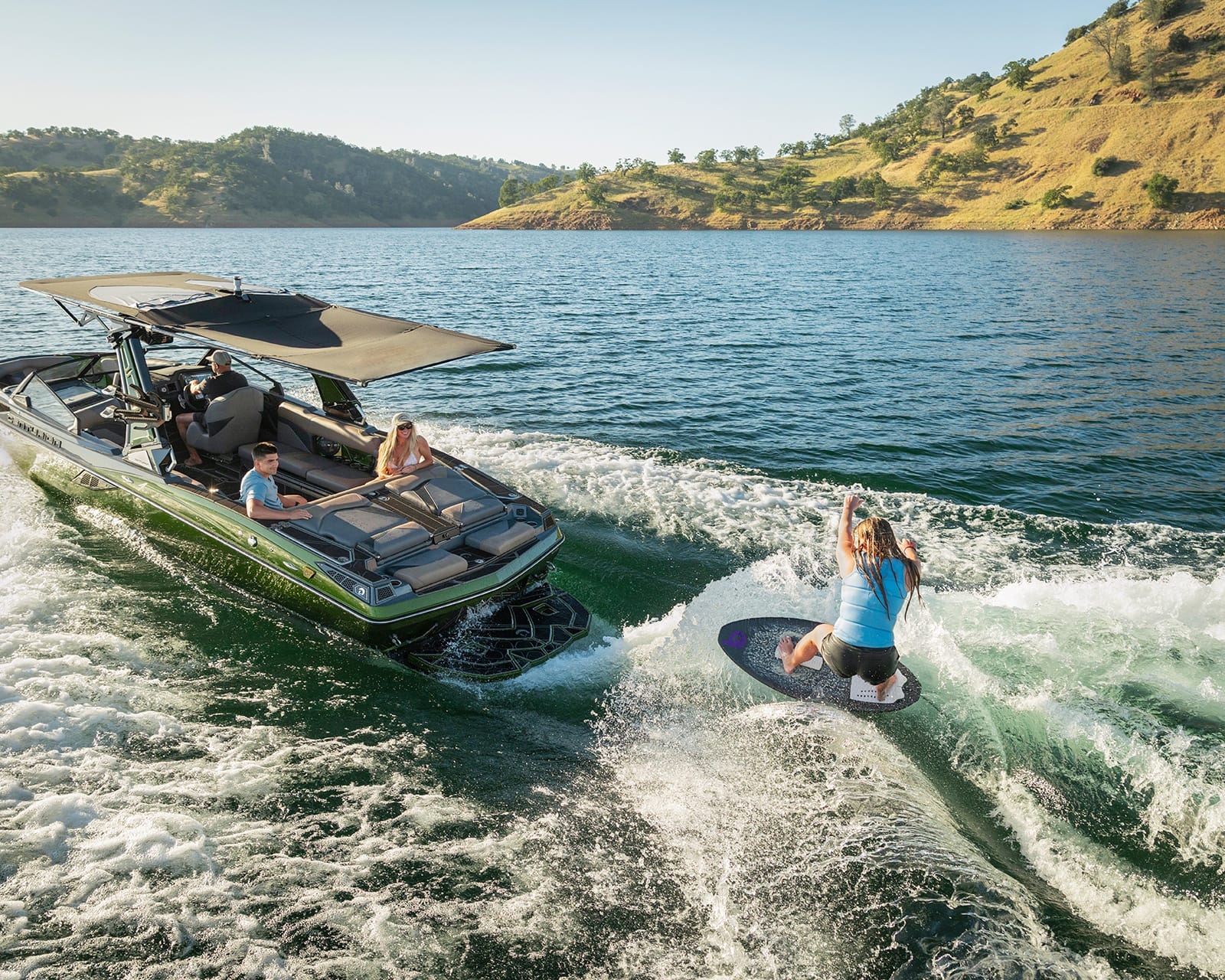 A person wake surfs behind a Centurion Fi25 on a lake, while two people watch from inside the boat near a hilly shoreline under a clear sky.