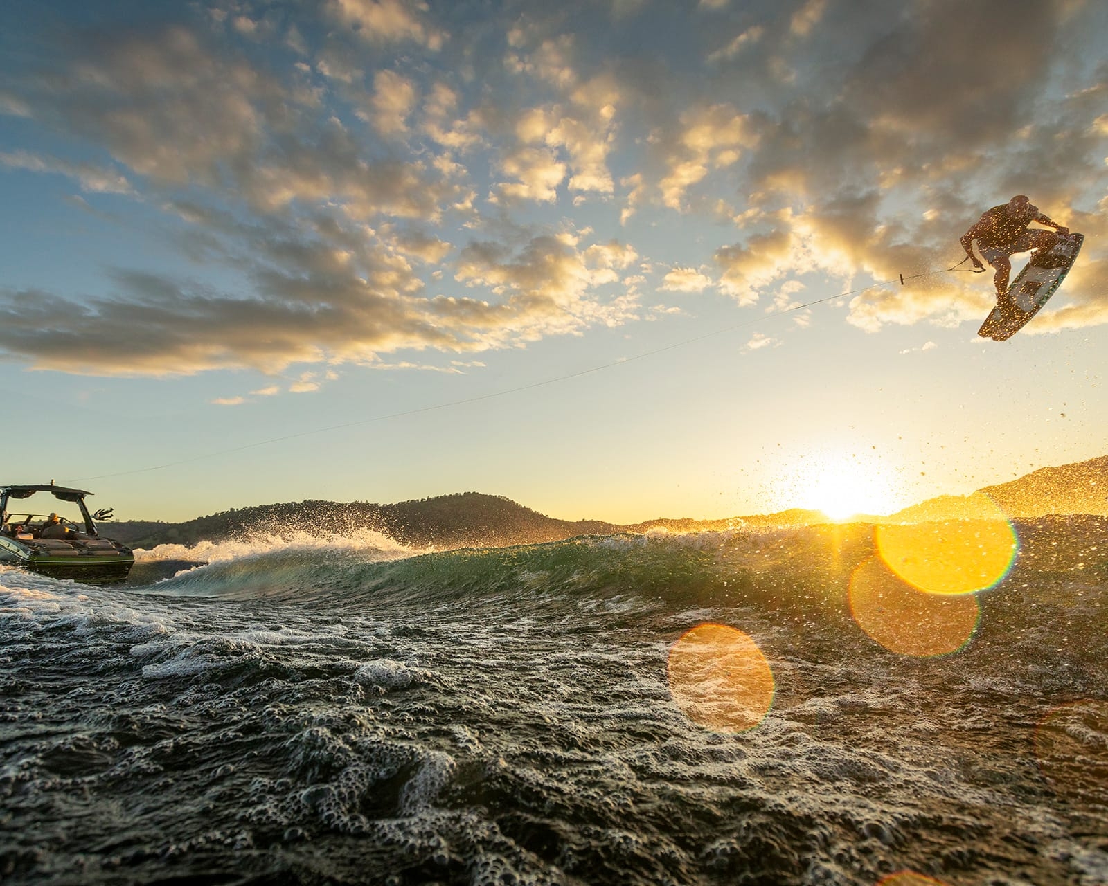 A person wakeboarding is airborne behind a Centurion Fi25 on a lake at sunset, with splashing water and hills in the background.