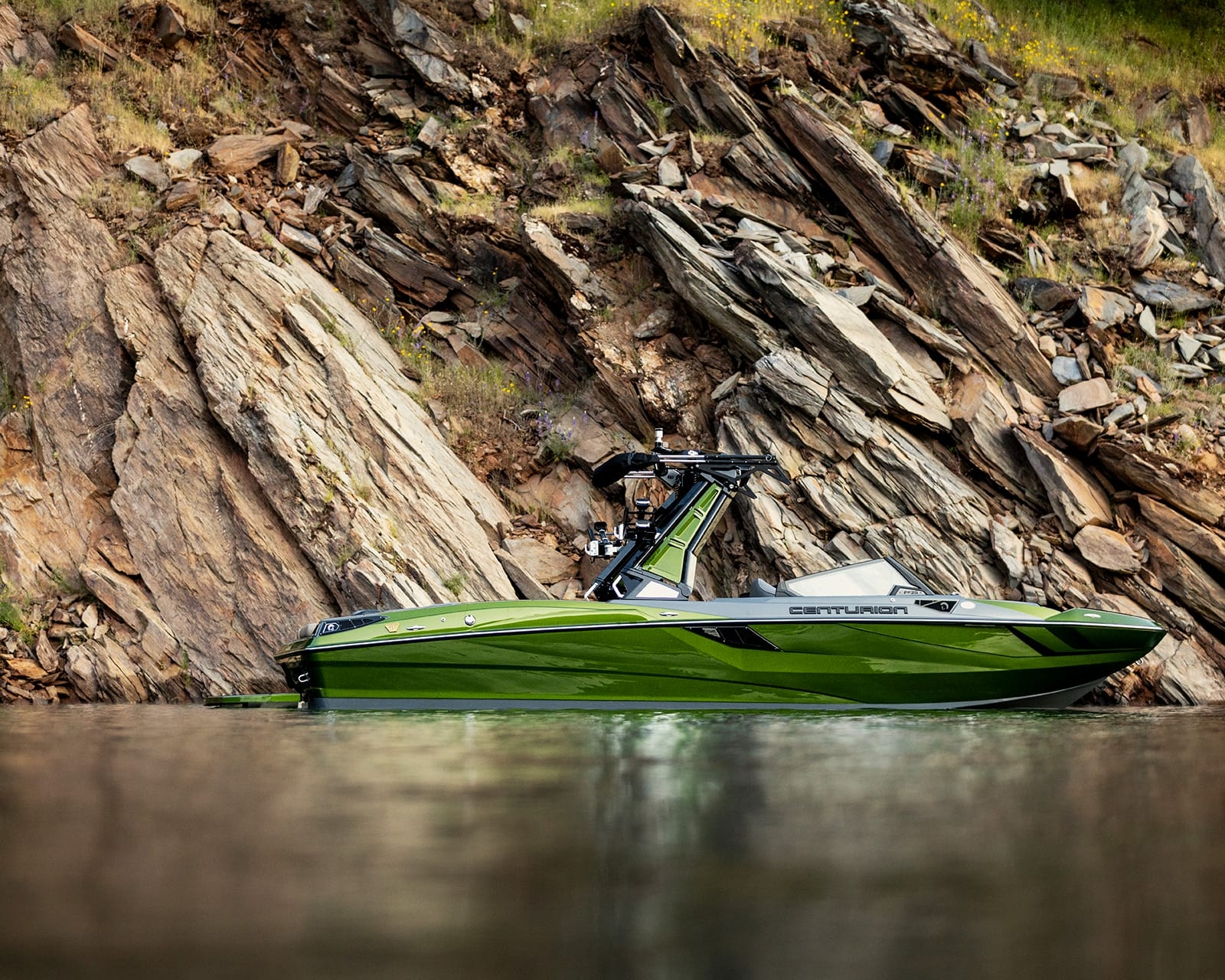 A green Centurion Fi25 Wake Boat is floating on calm water near a rocky shoreline with layered stone formations and sparse vegetation.