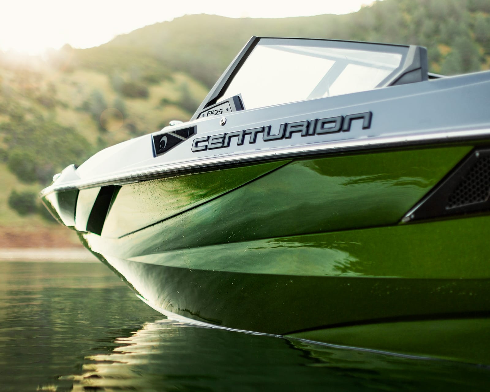 Close-up of a green Centurion Fi25 Wake Boat floating on calm water, with hills and trees in the background.