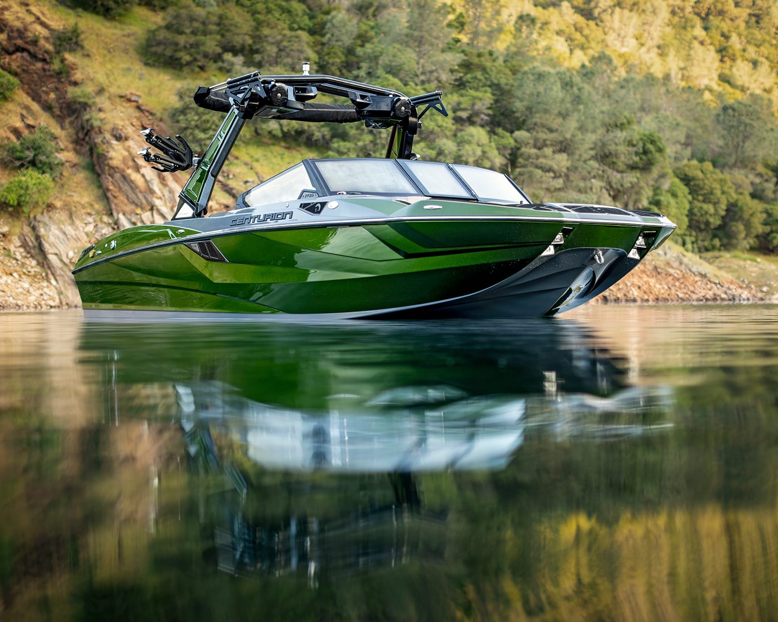 A green Centurion Fi25 with black accents floats on calm water near a rocky, tree-covered shoreline.