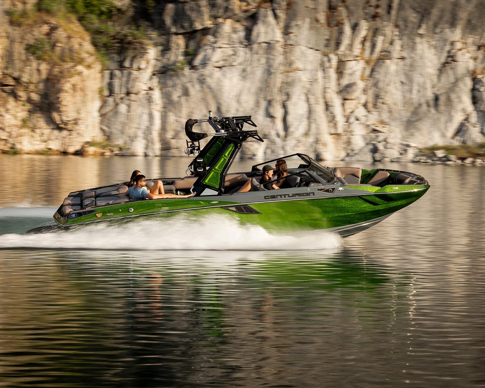 A green Centurion Fi25 speedboat with several people onboard moves quickly across a calm lake, rocky cliffs rising in the background.