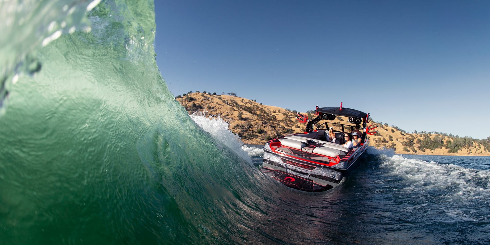 A Centurion Ri265 speedboat with people on board navigates on a clear blue water body under a sunny sky, creating a large wave beside the boat. Hills are visible in the background.