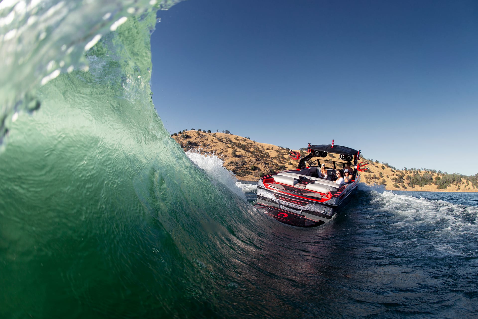 A Centurion Ri265 speedboat with people on board navigates on a clear blue water body under a sunny sky, creating a large wave beside the boat. Hills are visible in the background.