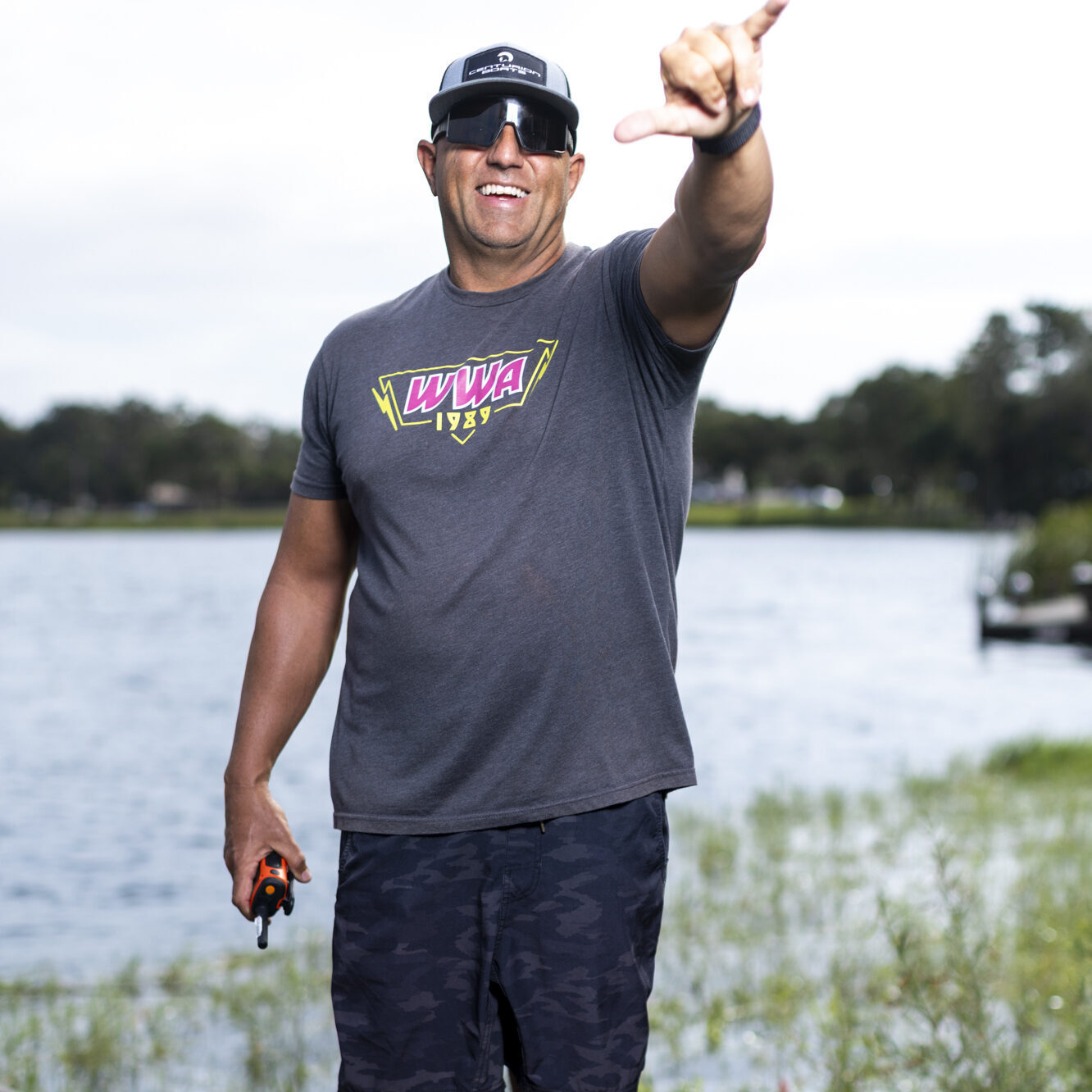 A man in sunglasses, a cap, and casual clothes stands on a dock by a lake, smiling and gesturing with his right hand, holding a small object in his left hand.