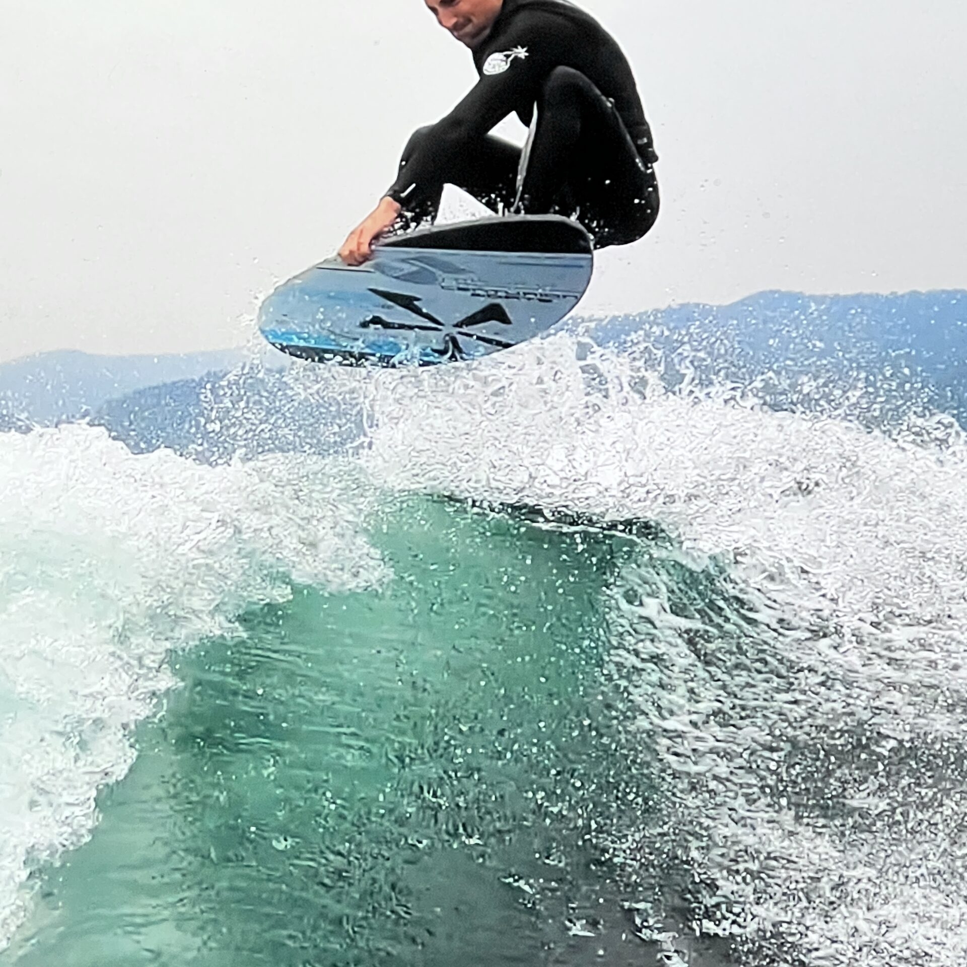 A man in a black wetsuit performs an aerial trick on a surfboard above a wave with water splashing around him.