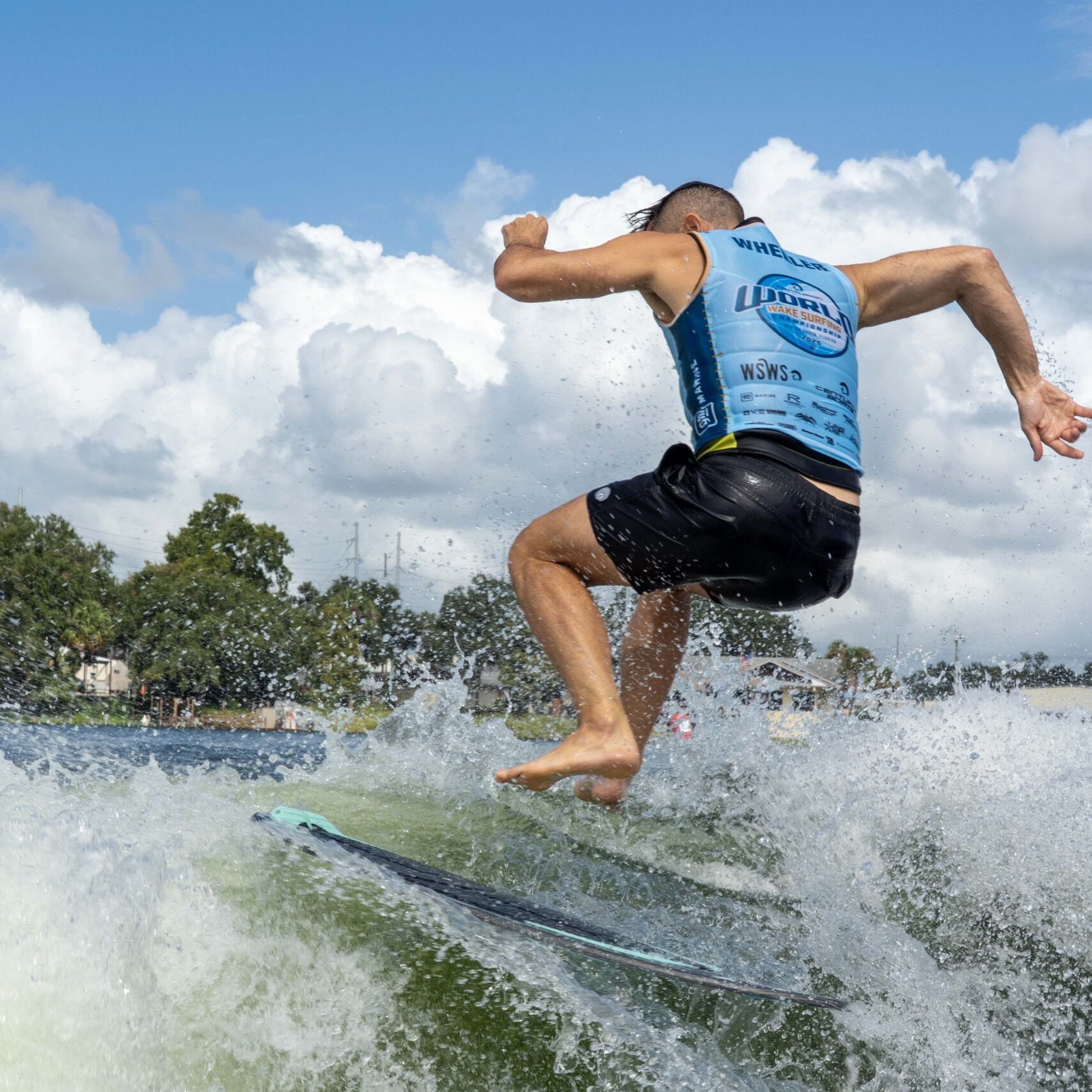 A man in a blue jersey and black shorts is wakesurfing, captured mid-air above the water with trees and buildings in the background.
