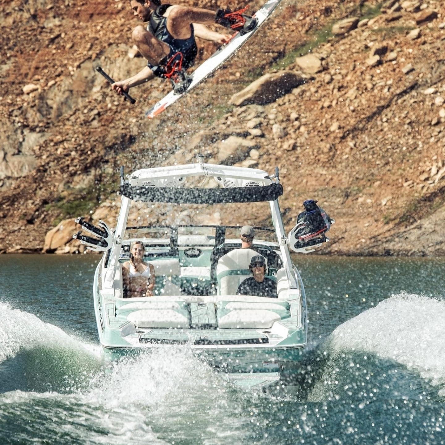 Person wakeboarding performs an airborne trick over a white and teal motorboat on a lake with rocky shores.