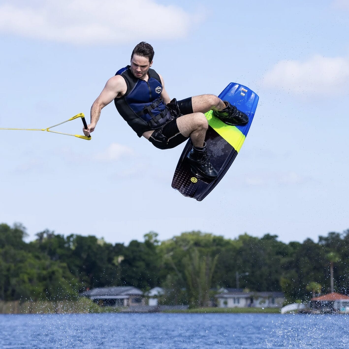 A person wakeboarding on a lake is captured mid-air, wearing a life jacket and holding a rope handle, with trees and houses in the background.