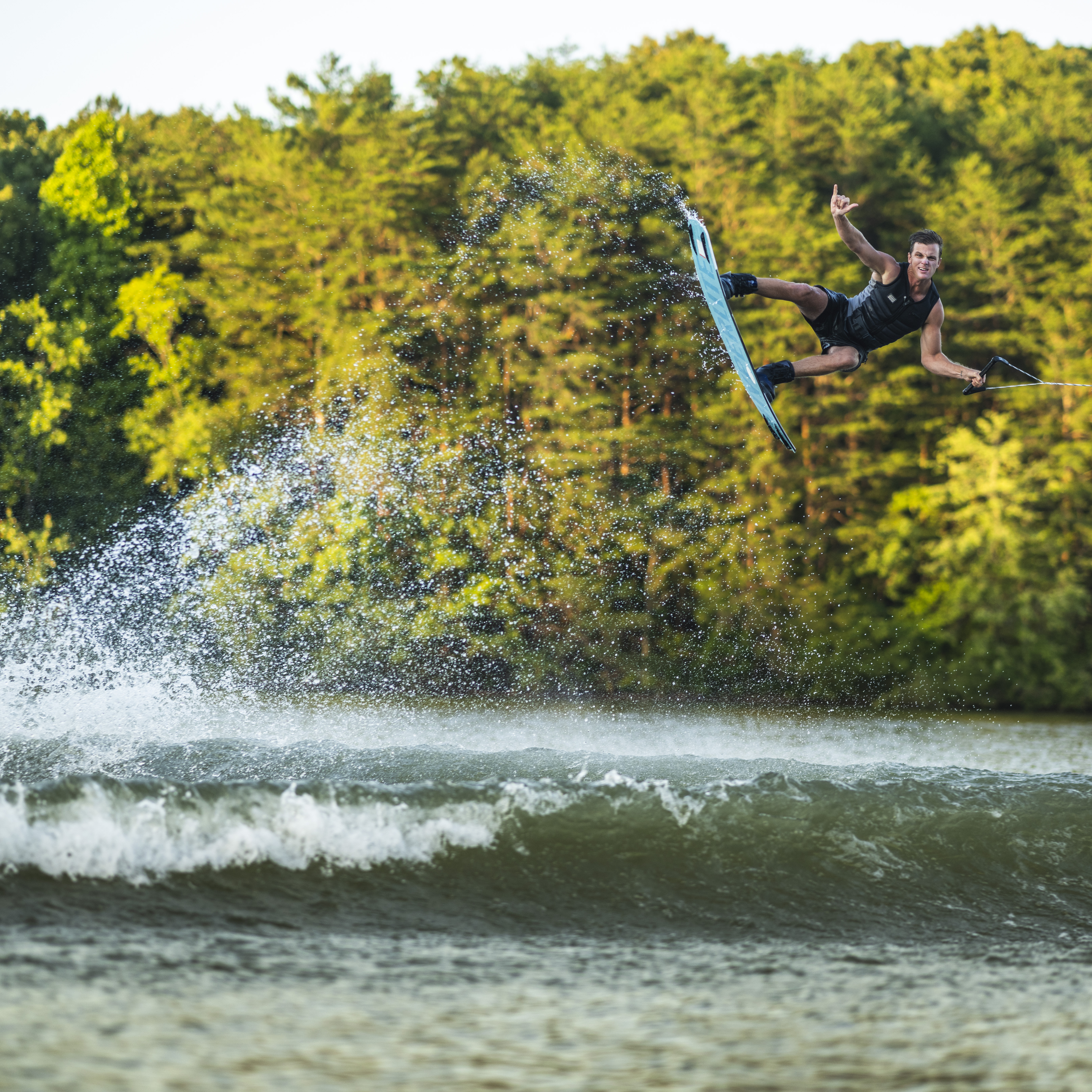 A person wakeboarding performs an aerial trick above a lake, with water splashing behind and dense green trees in the background.