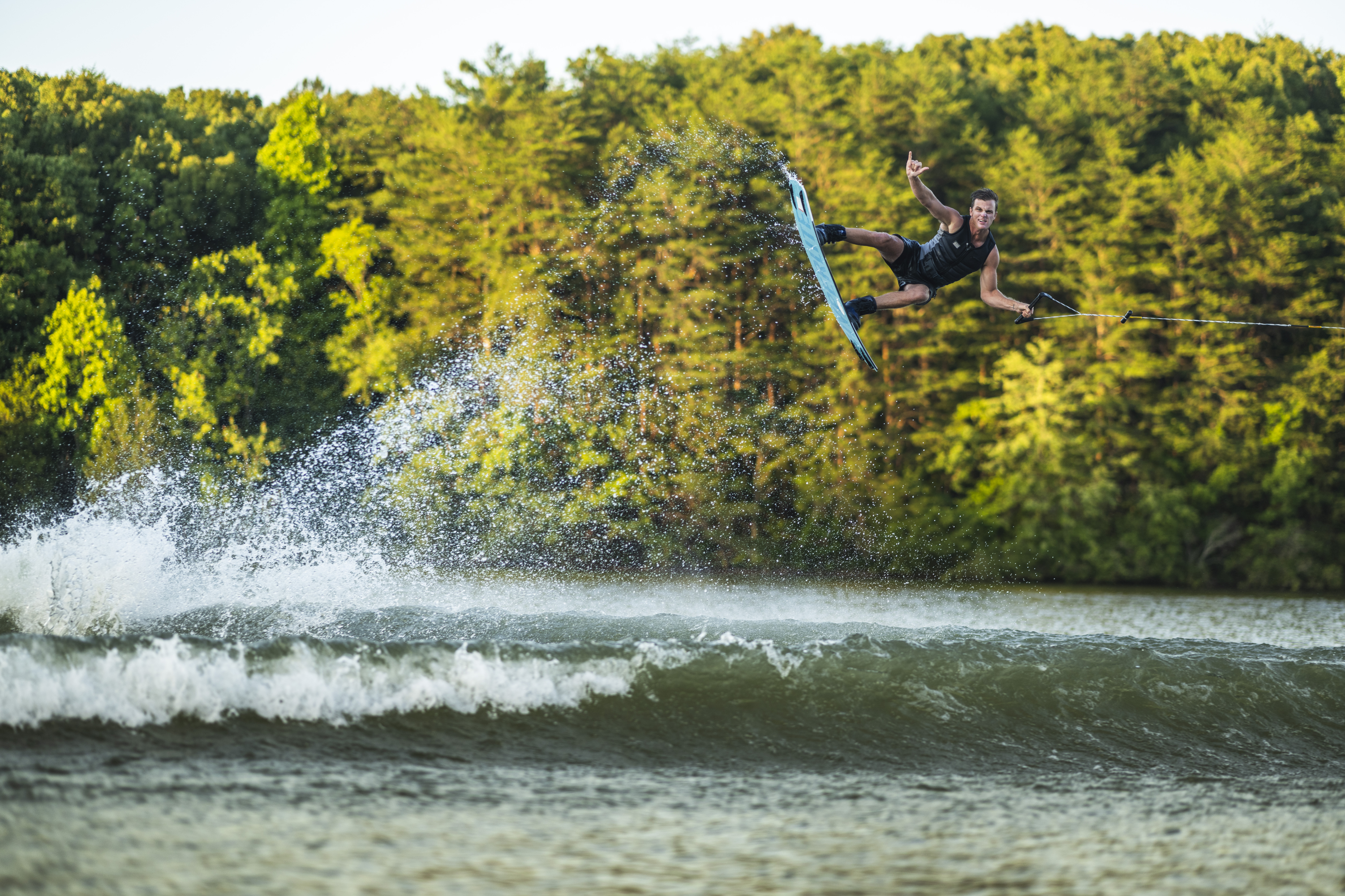A person wakeboarding performs an aerial trick above a lake, with water splashing behind and dense green trees in the background.