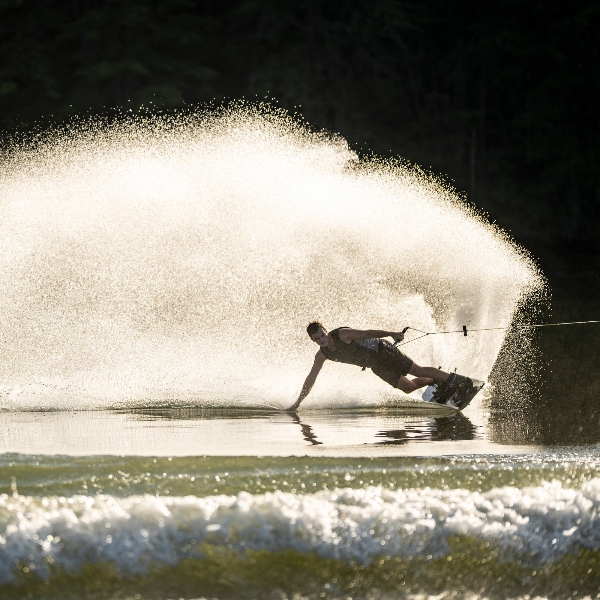 A person wakeboarding on a lake creates a large spray of water while leaning back and holding onto a tow rope.