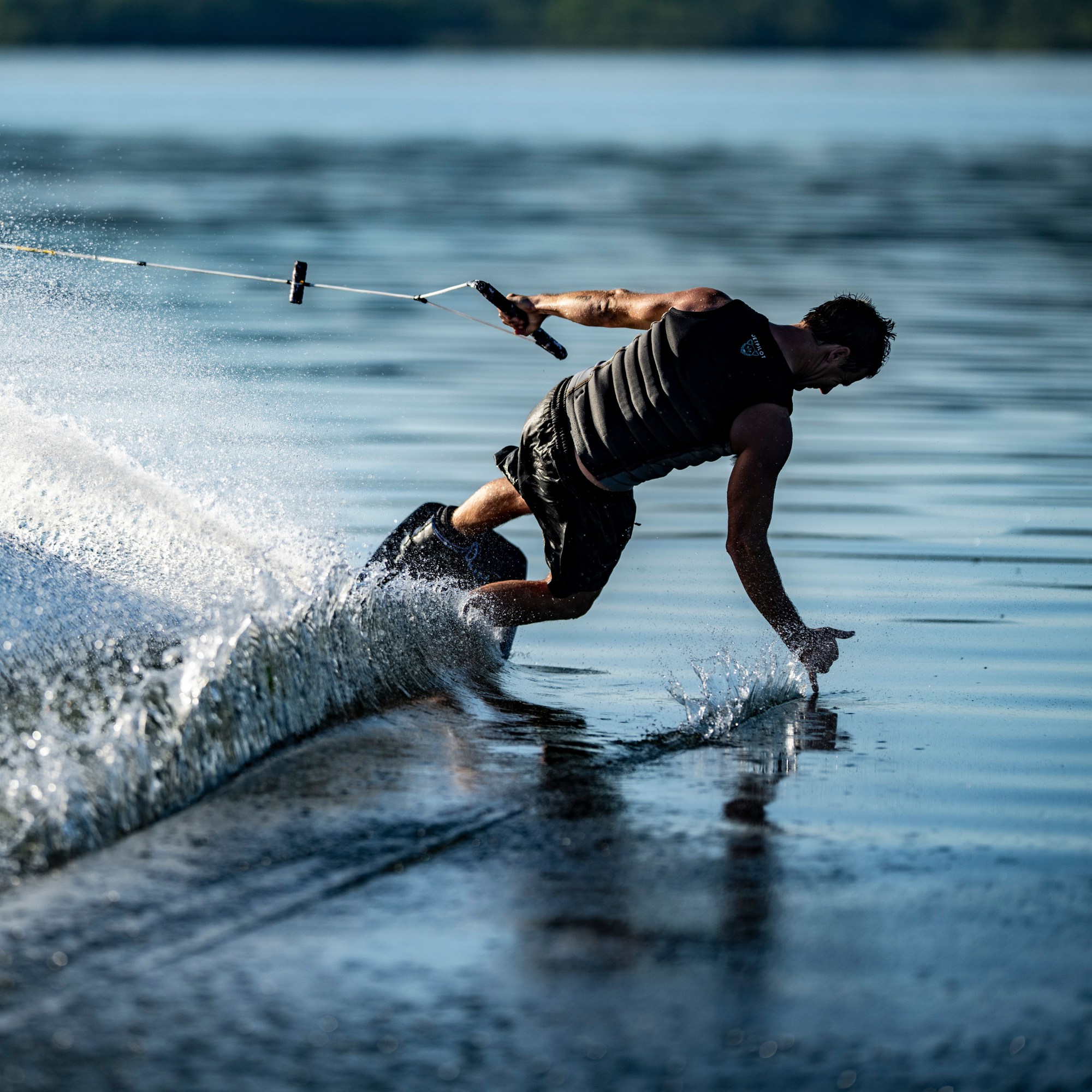 A person in a life vest is wakeboarding on a lake, leaning down with one hand touching the water while holding a tow rope with the other hand.