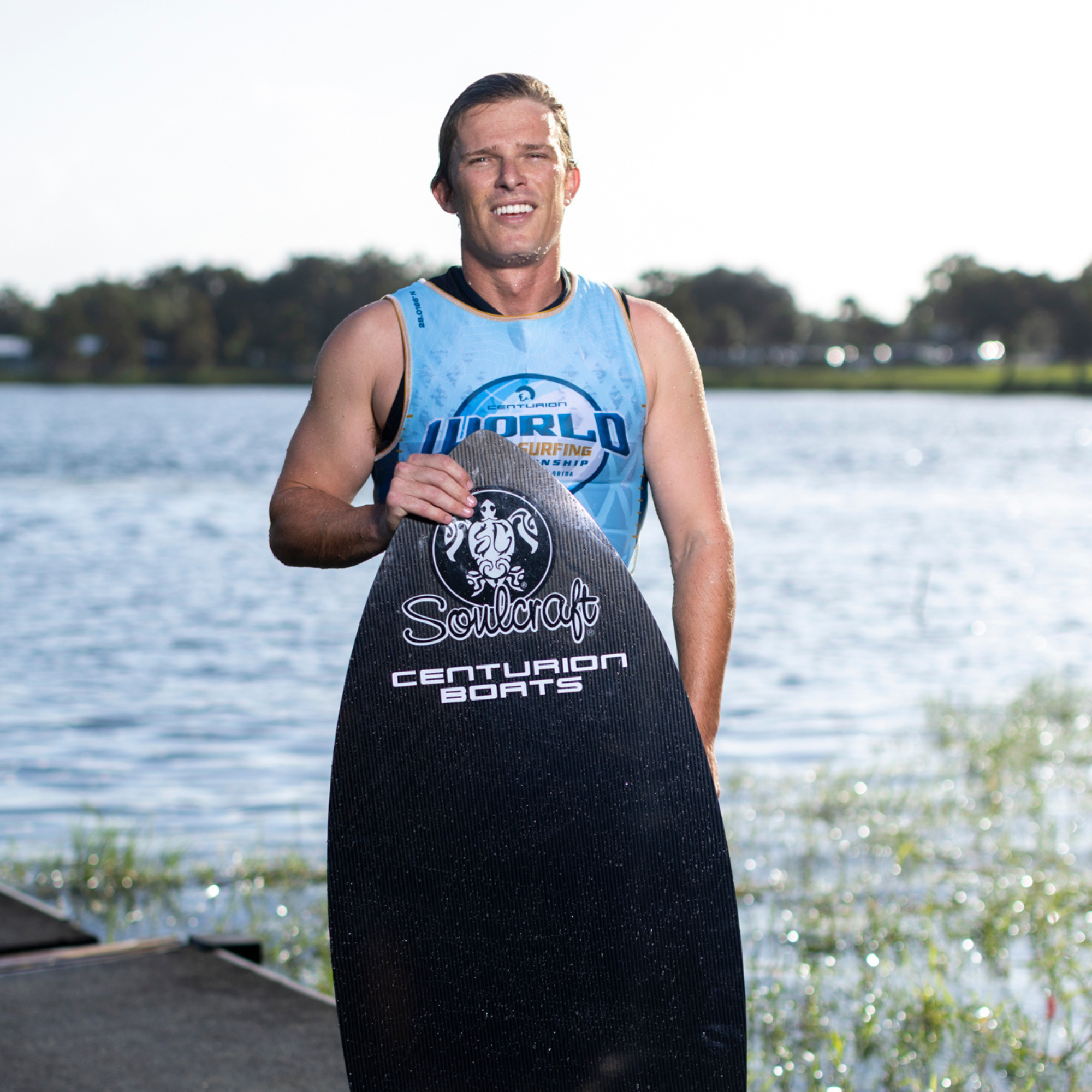 A man in a sleeveless shirt stands by a lake, holding a Soulcraft Centurion Boats wakesurf board and smiling at the camera.