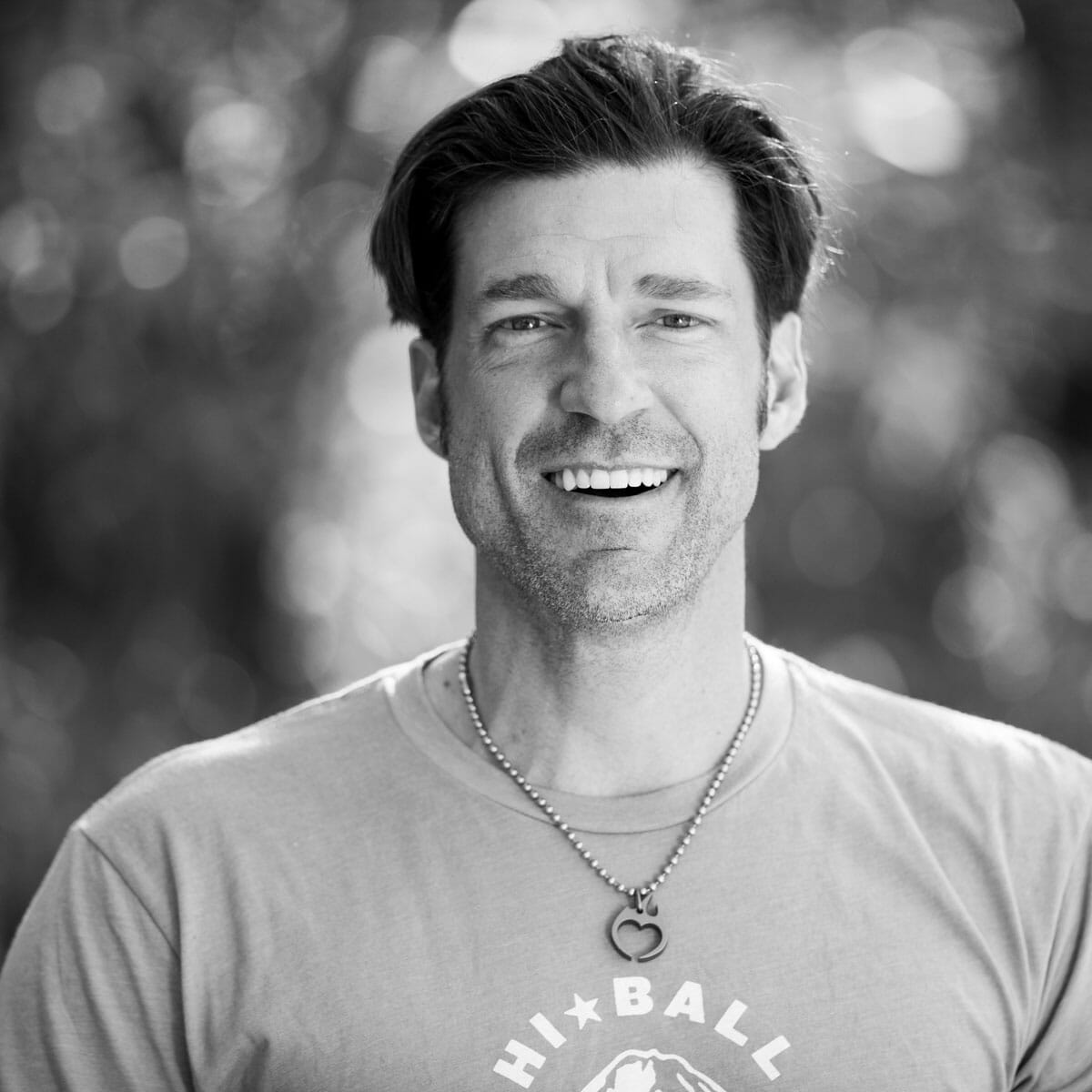 A black and white photo of a man smiling while wakesurfing behind a wakeboat.