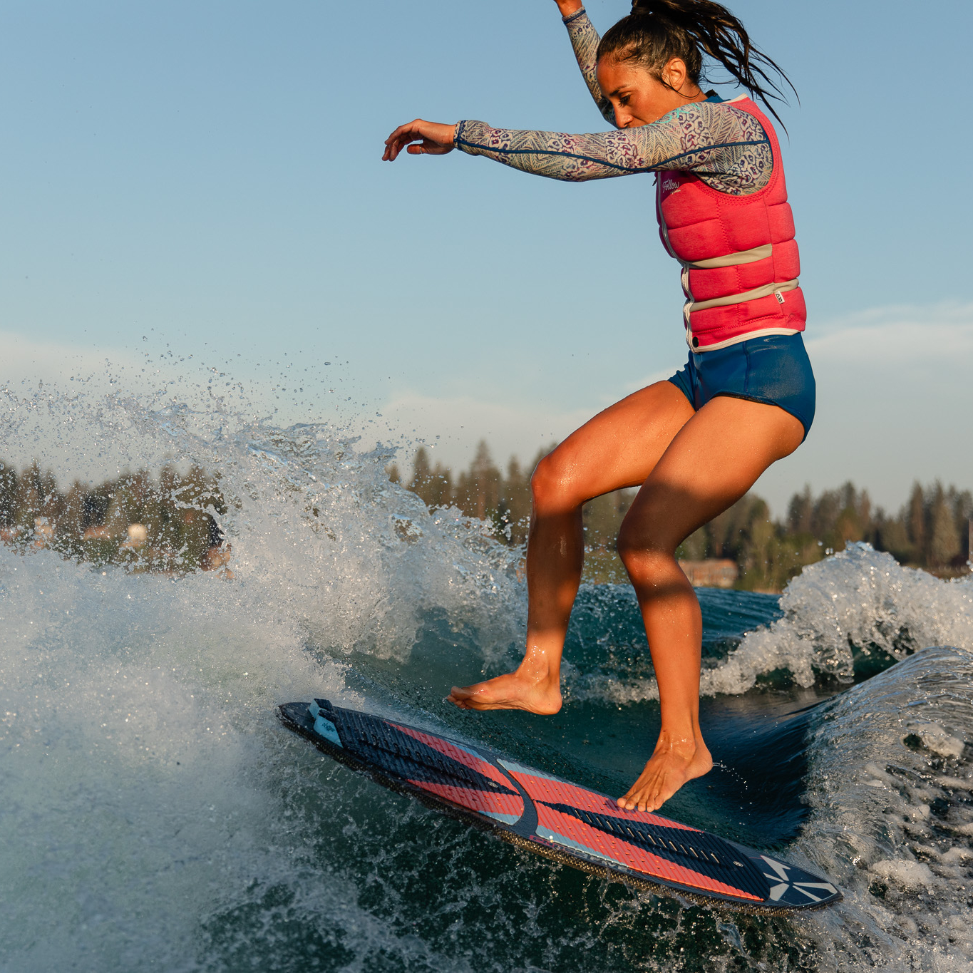A person wearing a pink life vest and patterned sleeves surfs on a wave, balancing on a surfboard with trees visible in the background.
