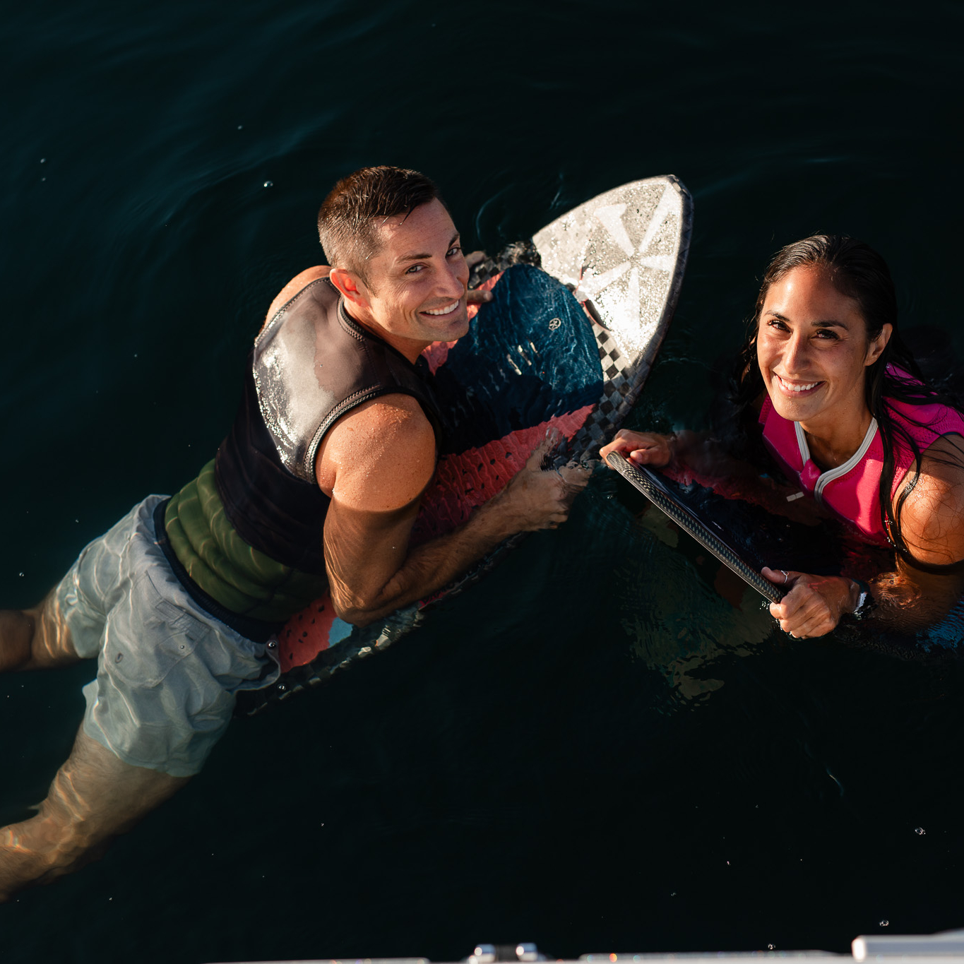 Two people wearing swim vests are floating in dark water, each holding onto a surfboard, and smiling up at the camera.