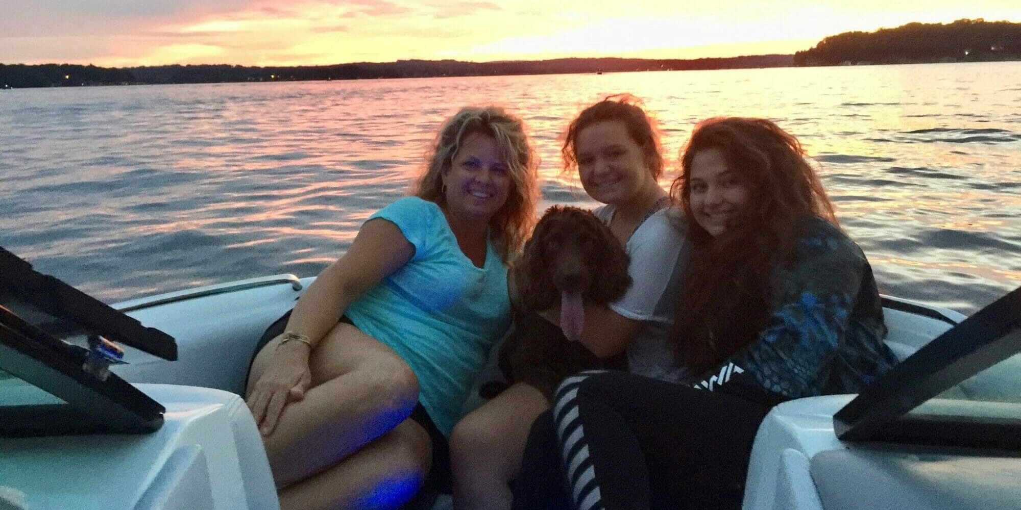 Three women and a dog on a wakesurf boat at sunset.