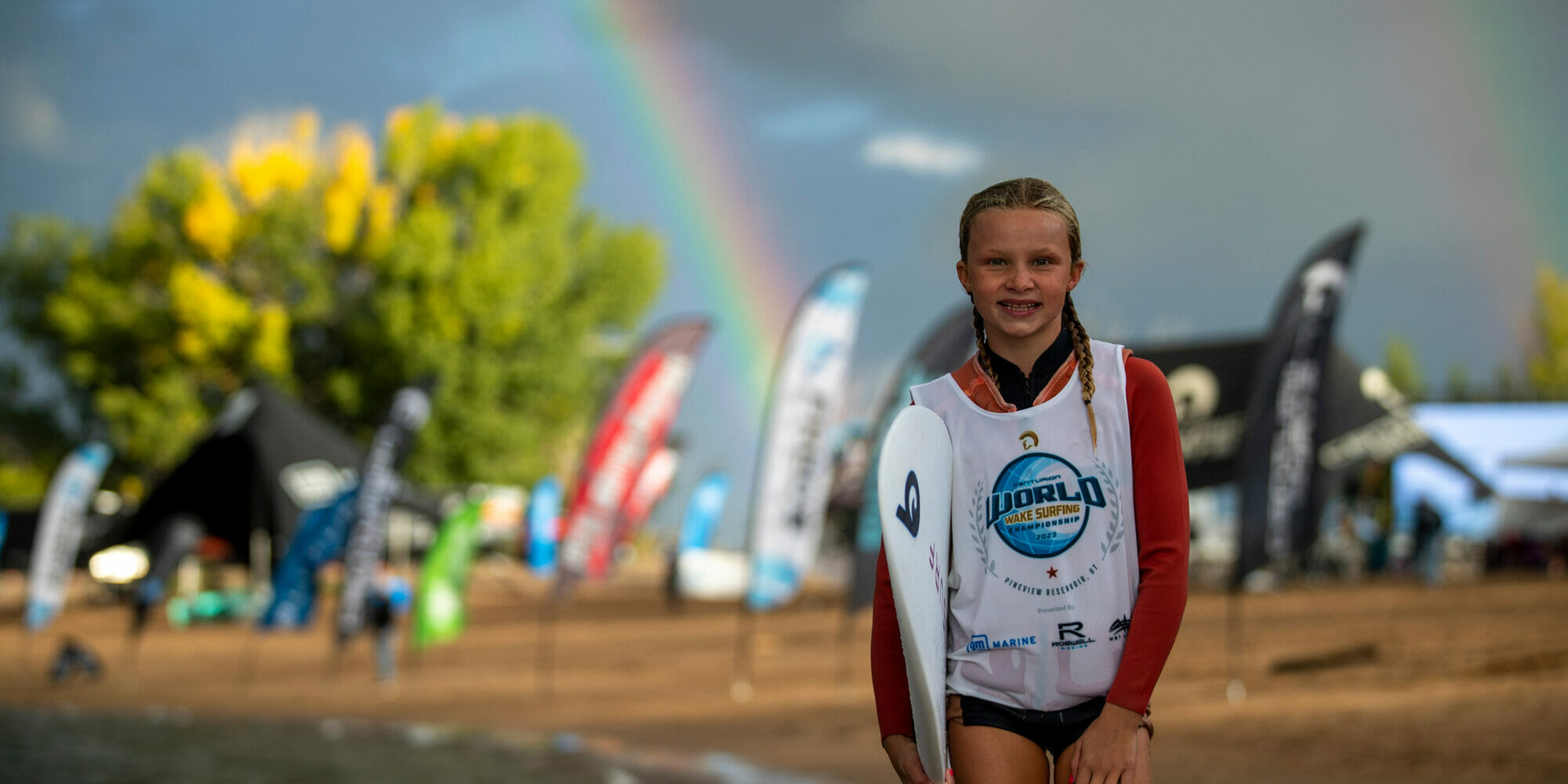 A girl with a wakesurf board standing on the beach with a rainbow in the background.