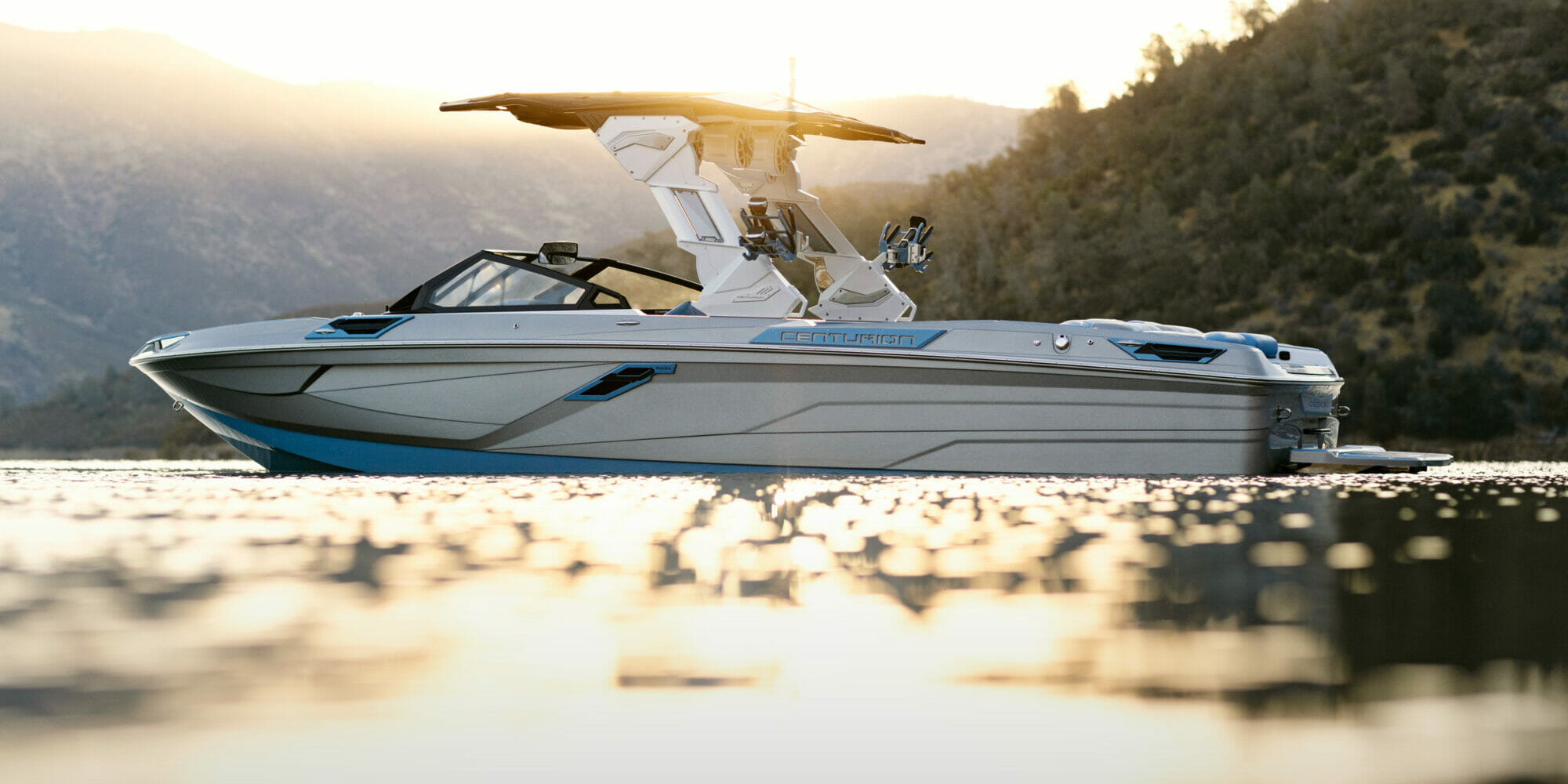 A wakeboat on the water with mountains in the background.