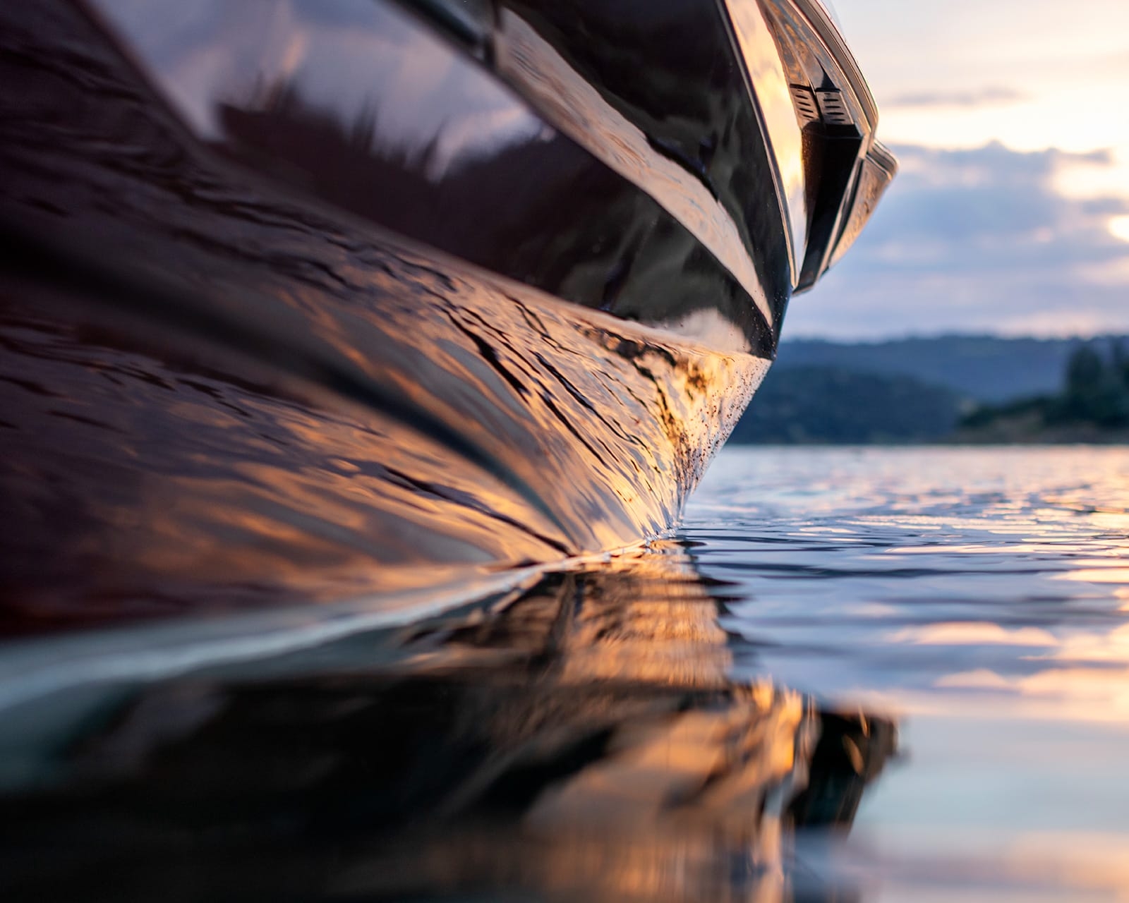 Close-up view of a Centurion Fe22 hull at water level, reflecting sunlight during sunset, with rippling water and a distant shoreline in the background.