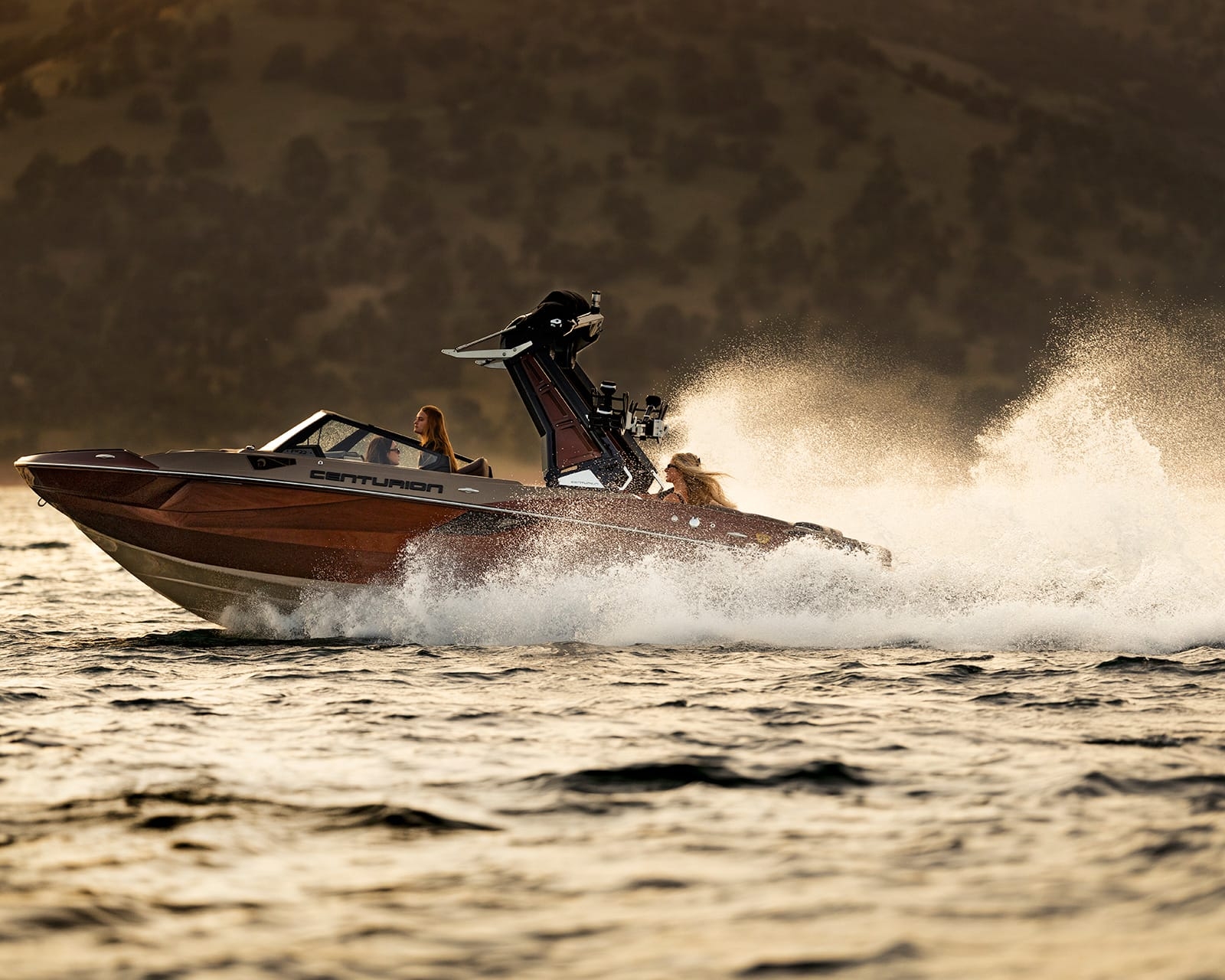 A Centurion Fe22 speeds across the lake with three people on board, creating splashes as it cuts through the water, framed by tree-covered hills in the background.