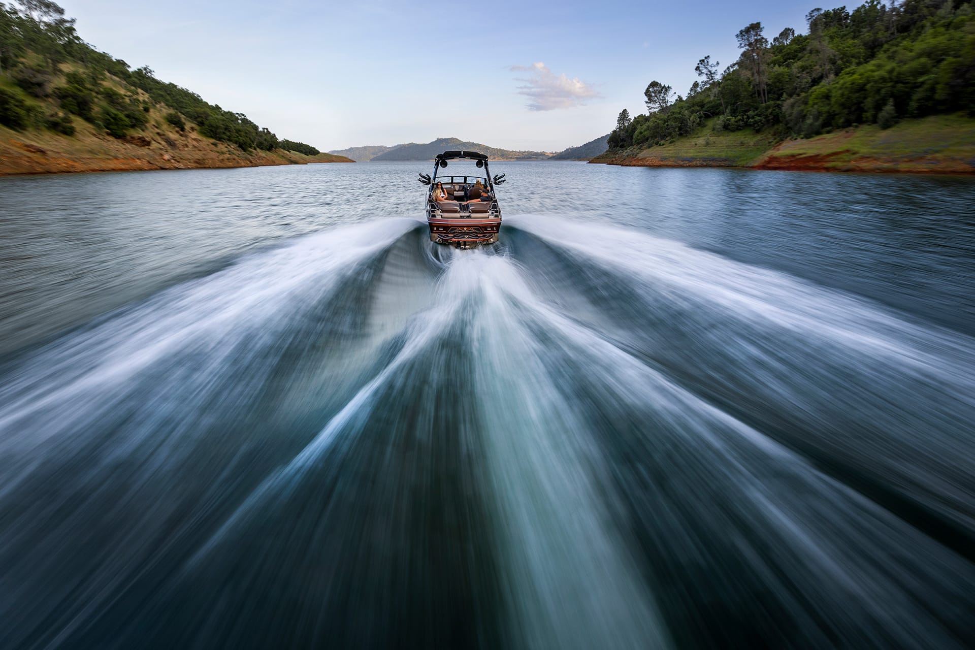 A Centurion Fe22 Wake Boat moves quickly across a wide lake, leaving a large wake behind, with forested hills on both sides and a partly cloudy sky overhead.
