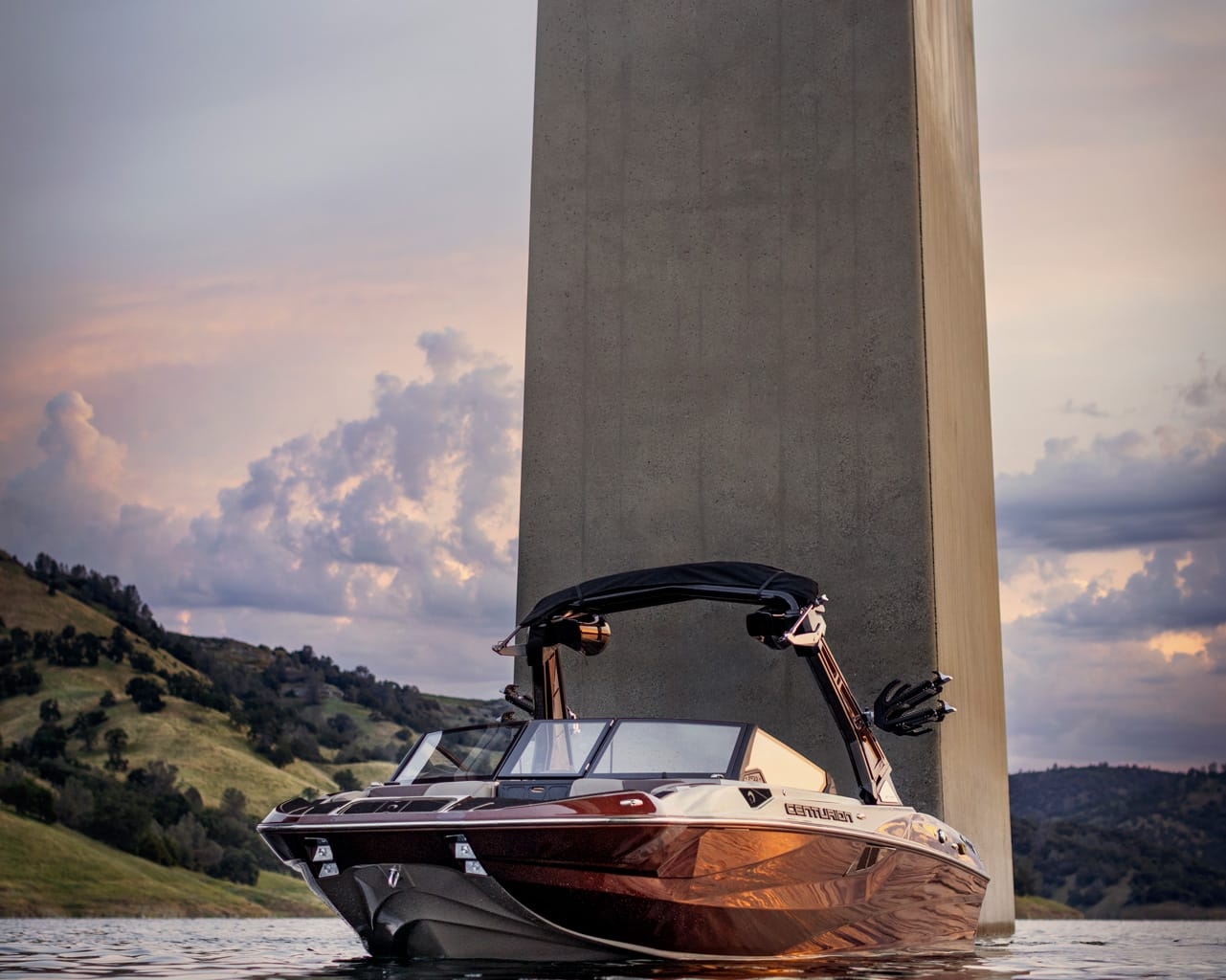 A Centurion Fe22 Wake Boat floats on calm water near the base of a large concrete bridge support, with hills and a cloudy sky in the background.