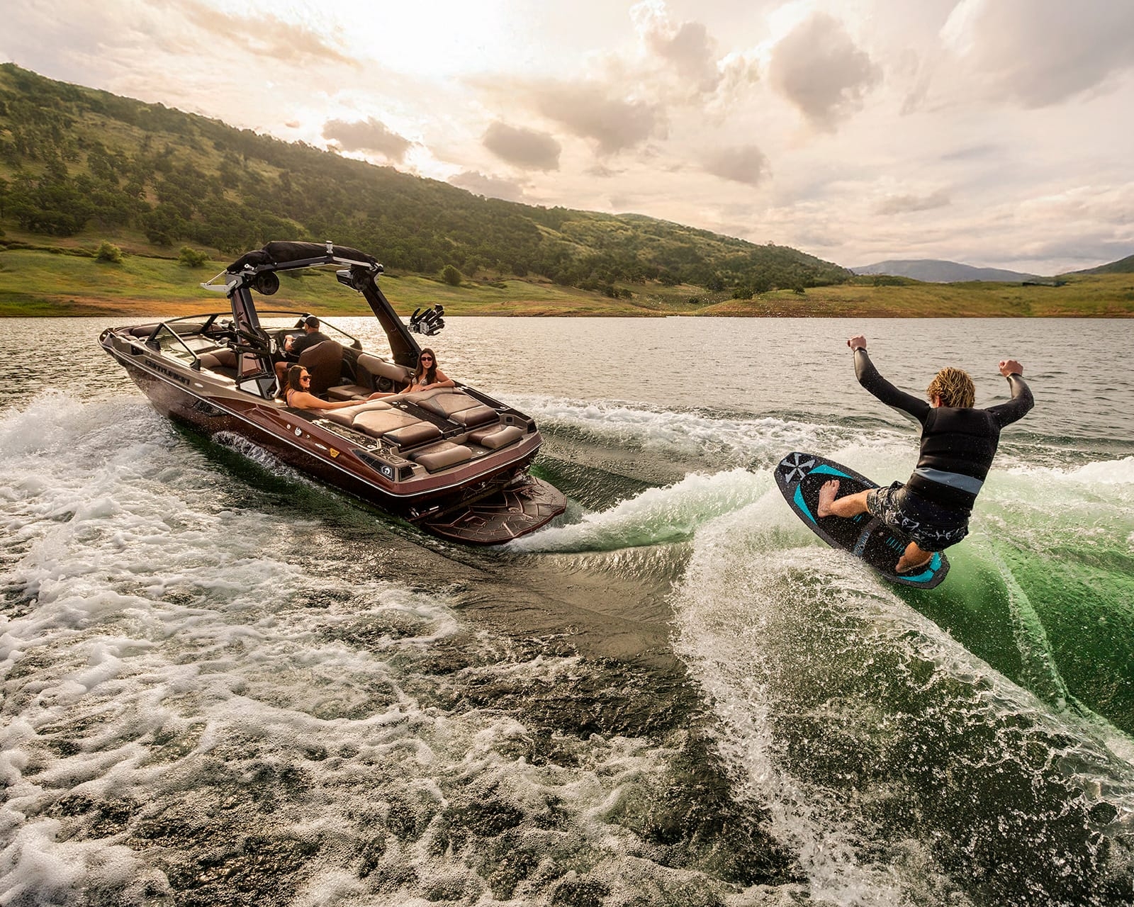 A person is wakesurfing behind a Centurion Fe22 Wake Boat on a lake surrounded by hills, with partly cloudy skies overhead.
