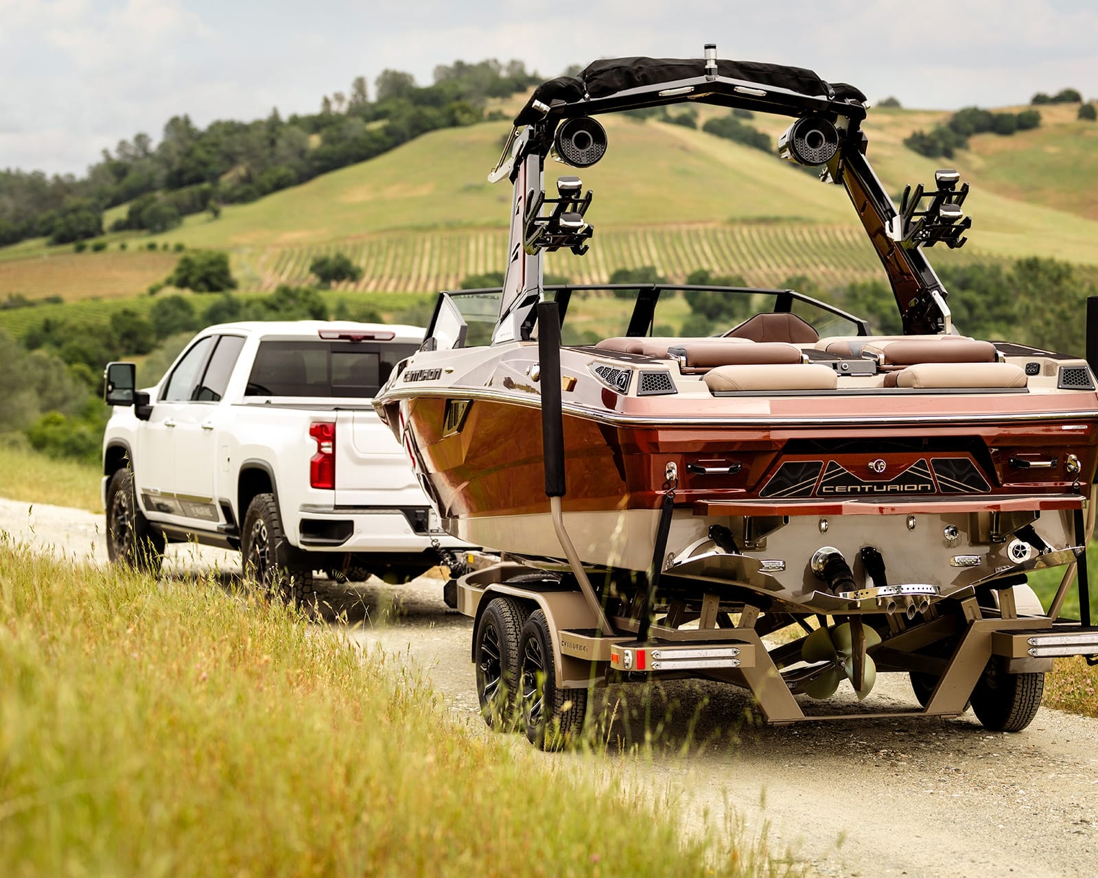 A white pickup truck is towing a brown and black Centurion Fe22 Surf Boat on a trailer down a gravel road, with green fields and hills in the background.