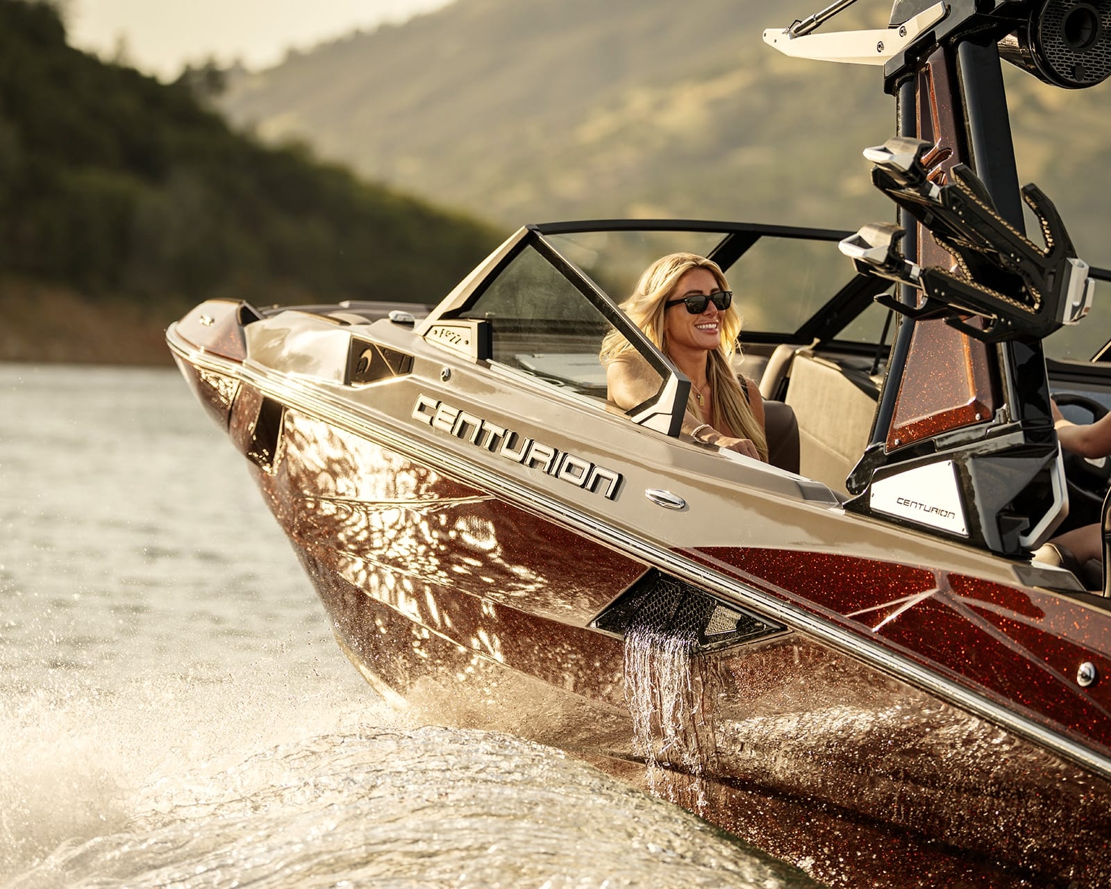 A woman wearing sunglasses smiles while sitting at the helm of a Centurion Fe22 Wake Boat cruising on a lake, with water splashing beside the boat.