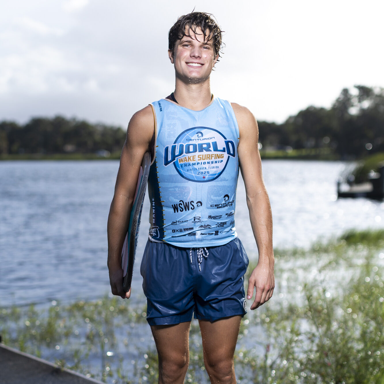 Luke Hutcherson stands barefoot on a dock by a lake, wearing a blue wake surfing tank top and shorts, holding a wakesurf board.