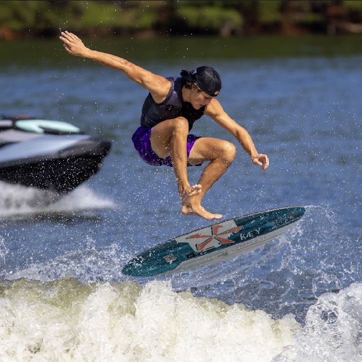 Luke Hutcherson, in a backwards cap and life vest, performs a trick on a wakeboard above the water, with a boat in the background.