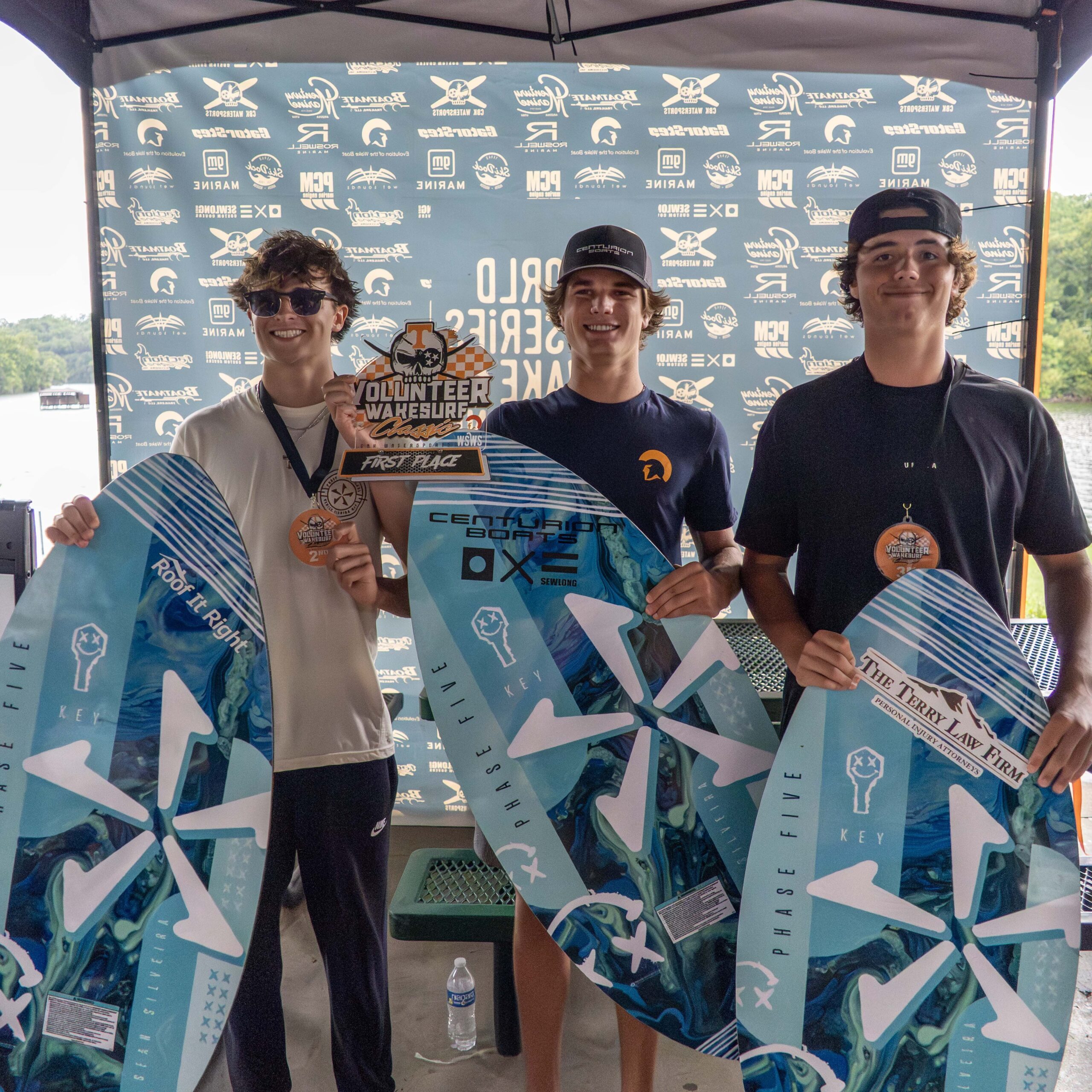 Three young men, including Luke Hutcherson, stand side by side holding wakeboards and medals in front of a branded backdrop at a wakeboard competition event.