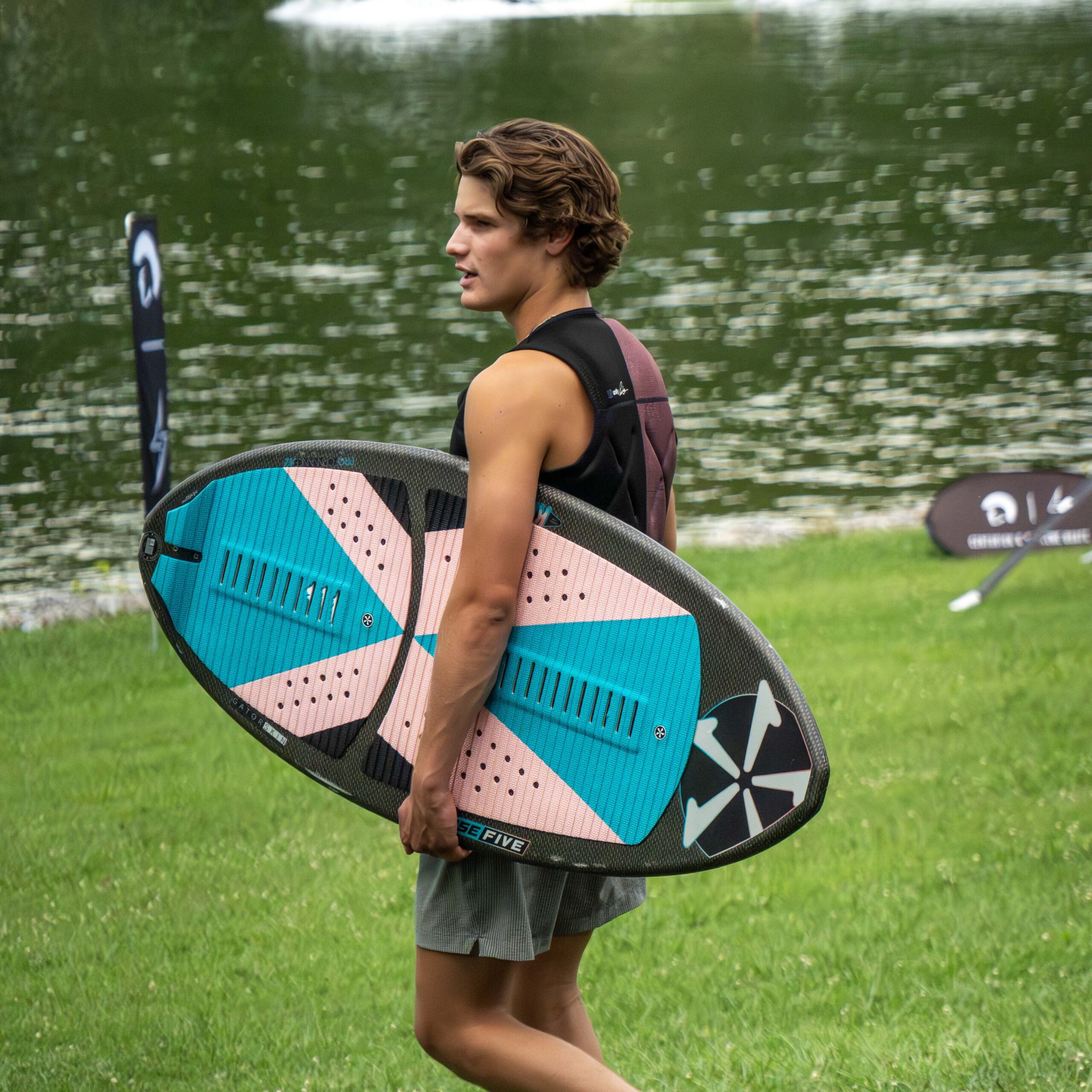 A person stands on grass near water, holding a colorful surfboard under one arm and wearing a sleeveless shirt and shorts, reminiscent of Luke Hutcherson’s laid-back style.