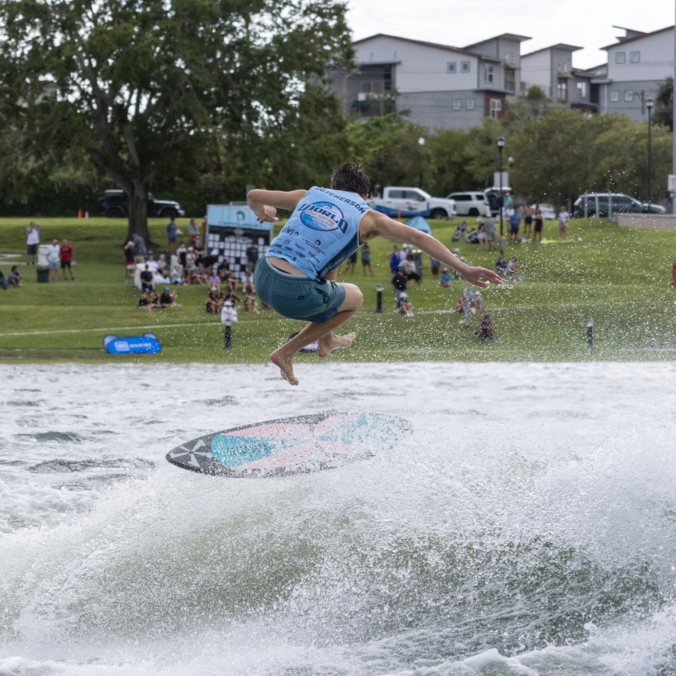 Luke Hutcherson, in a blue vest, is performing a trick on a wakeboard above the water, with spectators and parked cars in the background.