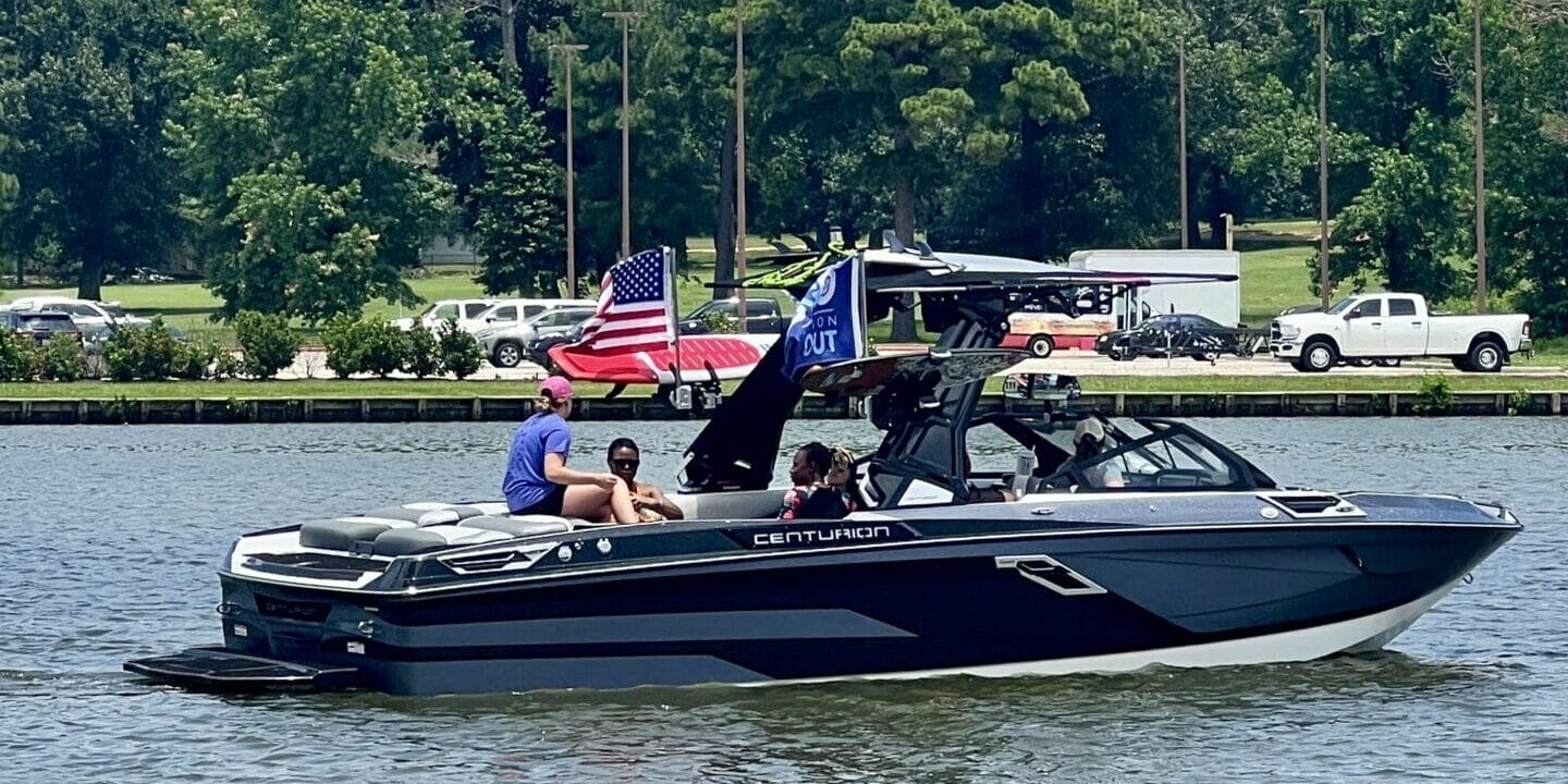 A Centurion Boats Salute to Service event honors heroes at the Come and Take it Wake Surf Classic in Lake Conroe, where a motorboat with several people on board navigates a calm lake. An American flag is visible on the boat, and trees and parked vehicles line the shoreline in the background.